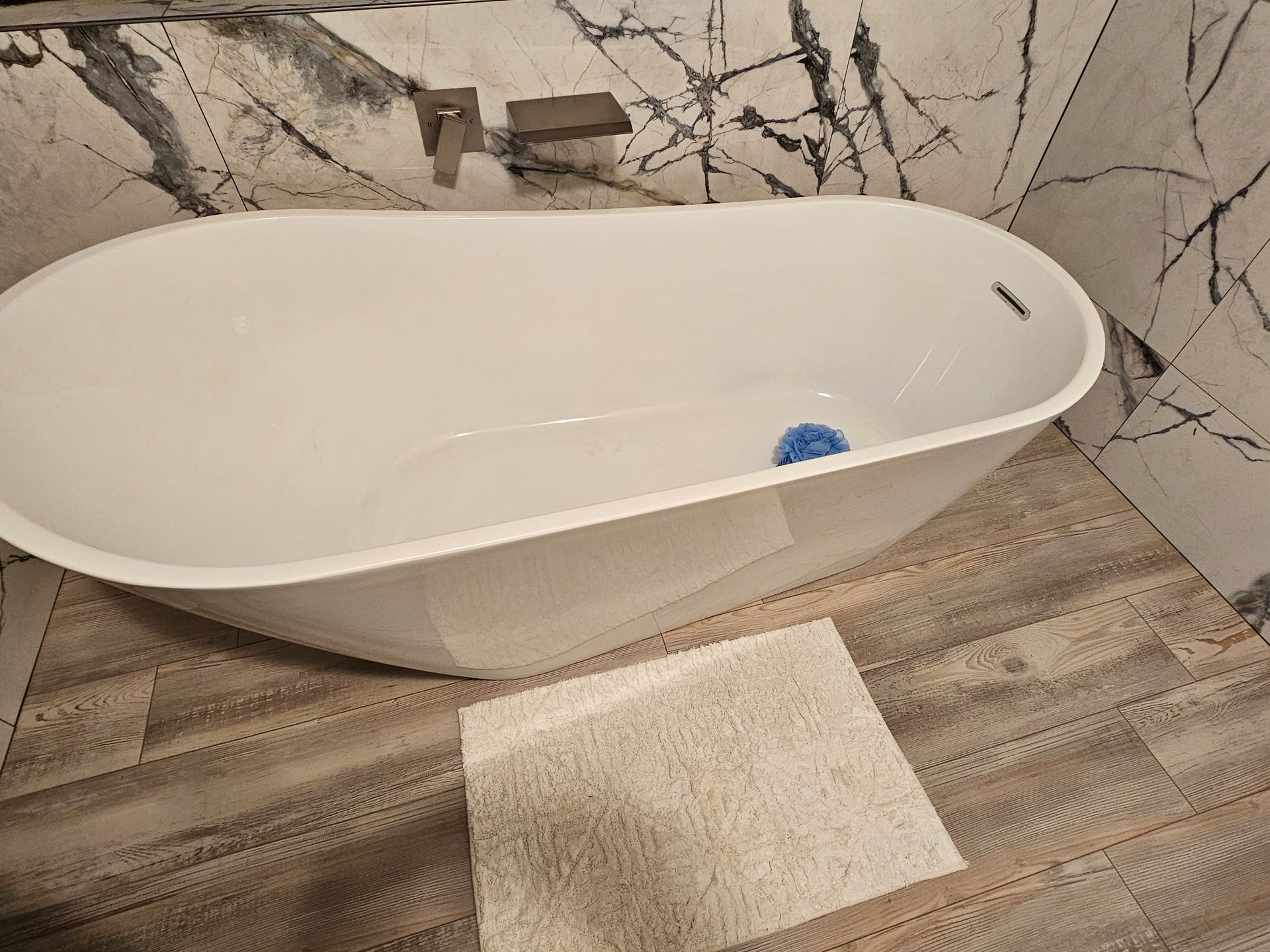 White freestanding bathtub in a marble-tiled bathroom with wood-look floor and a bath mat