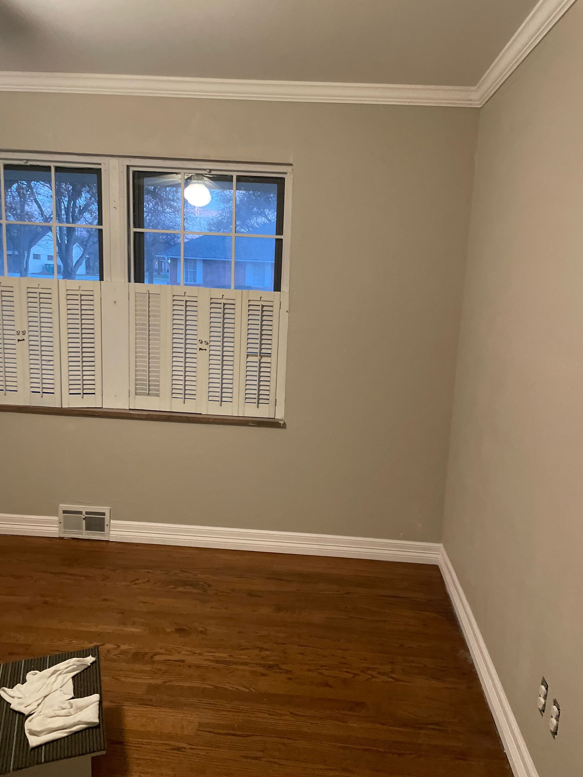 Empty beige room with hardwood floor, small window with white blinds, and one wall outlet.