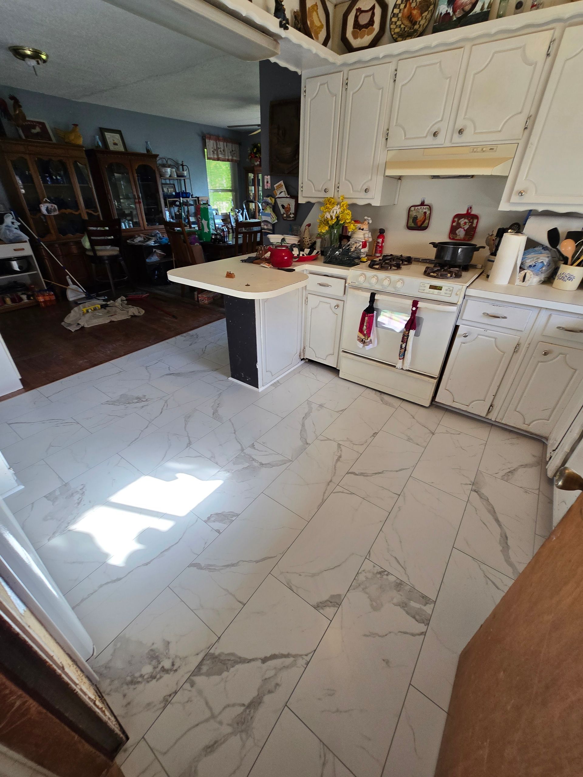 Bright kitchen with white cabinets, tiled floor, and a central island lit by sunlight, opening to a dining area