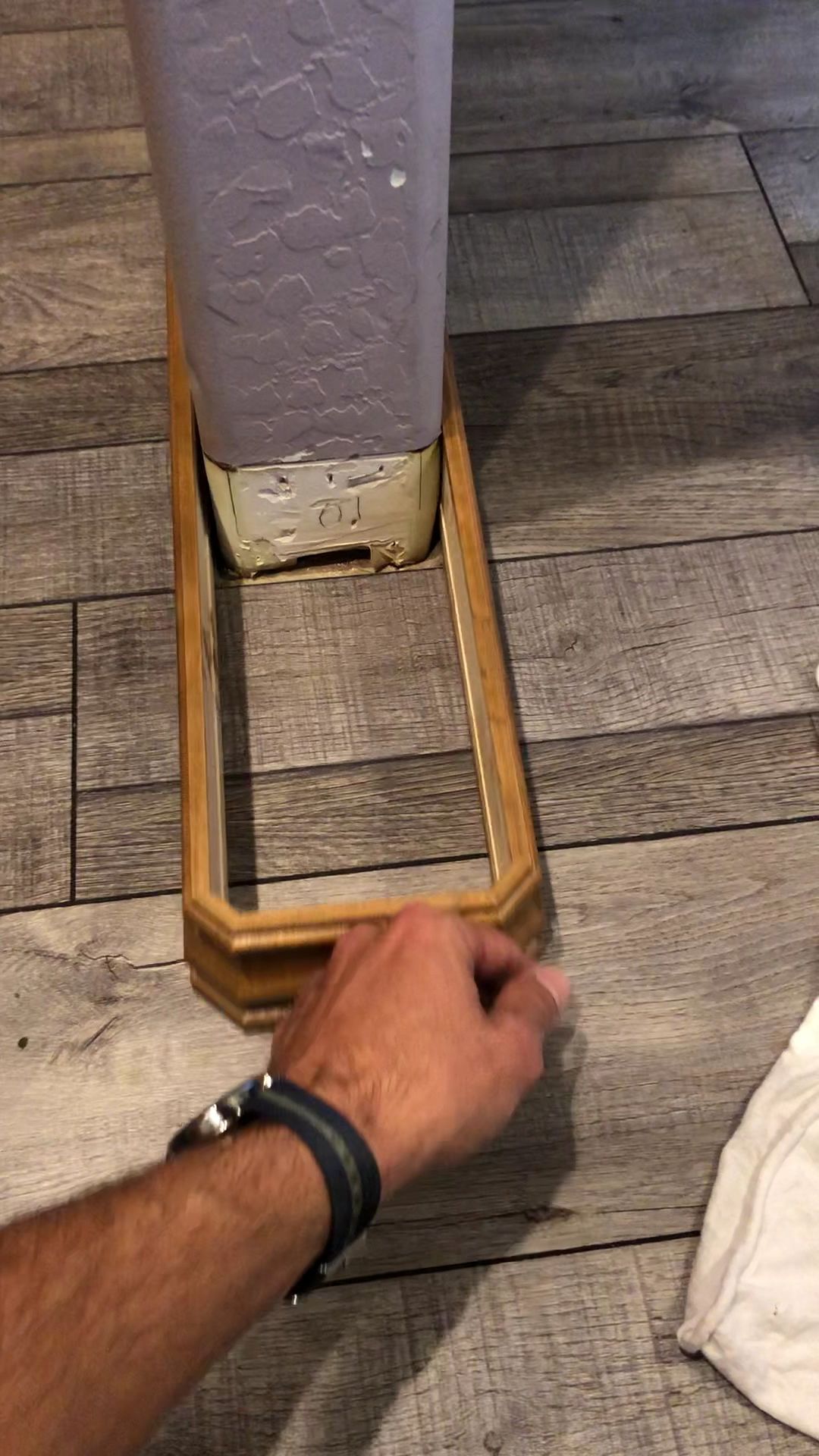 Hand measuring a floor gap beside a purple column with a wooden trim piece and tile floor