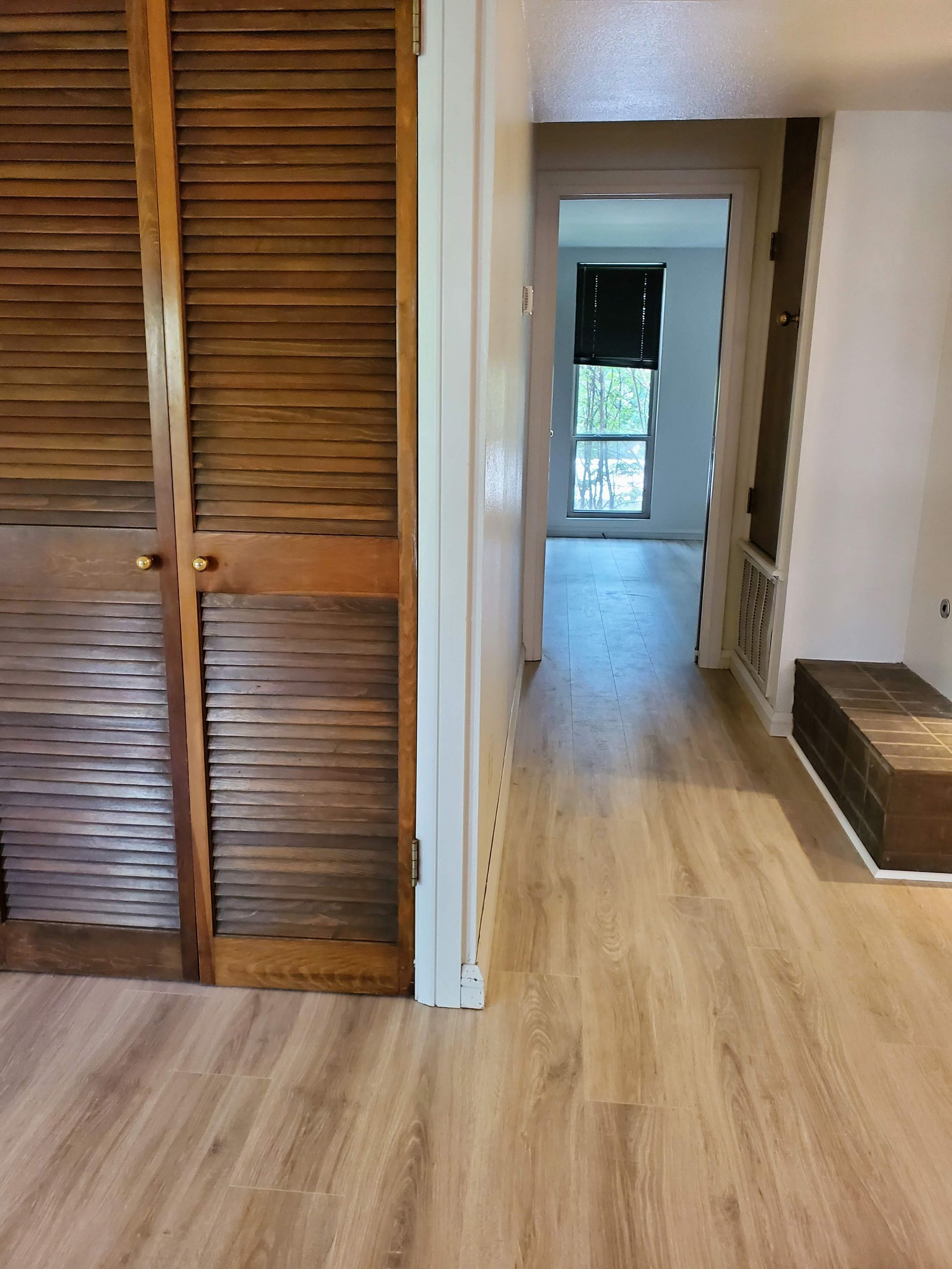 Interior hallway with wood floors, louvered closet doors, and a doorway leading to a bright room.