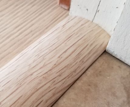 Light wood floorboard meeting a white baseboard and doorway trim on a beige floor edge