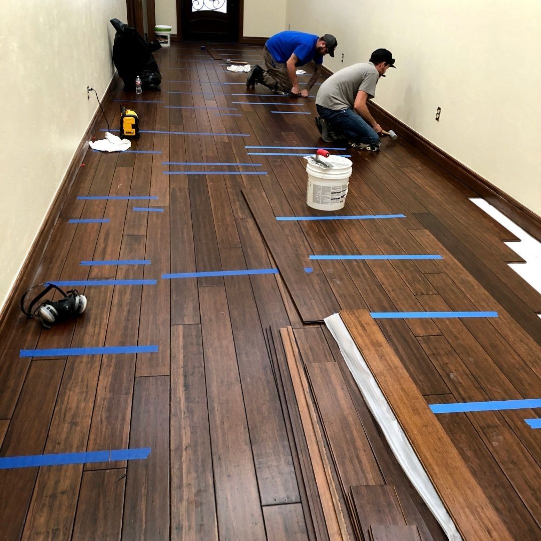 Workers installing blue tape on a wood floor in a long hallway, with tools and a white bucket nearby