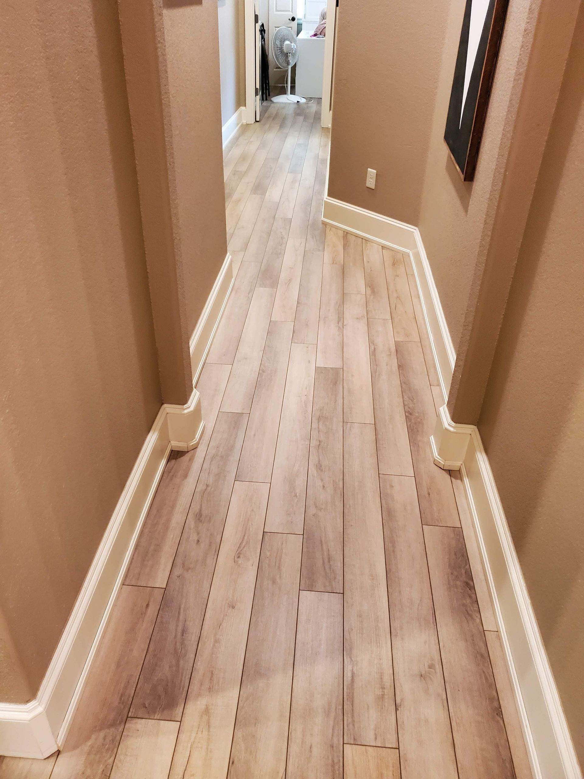 Narrow beige hallway with light wood floors and white trim, leading to a bright room at the end.
