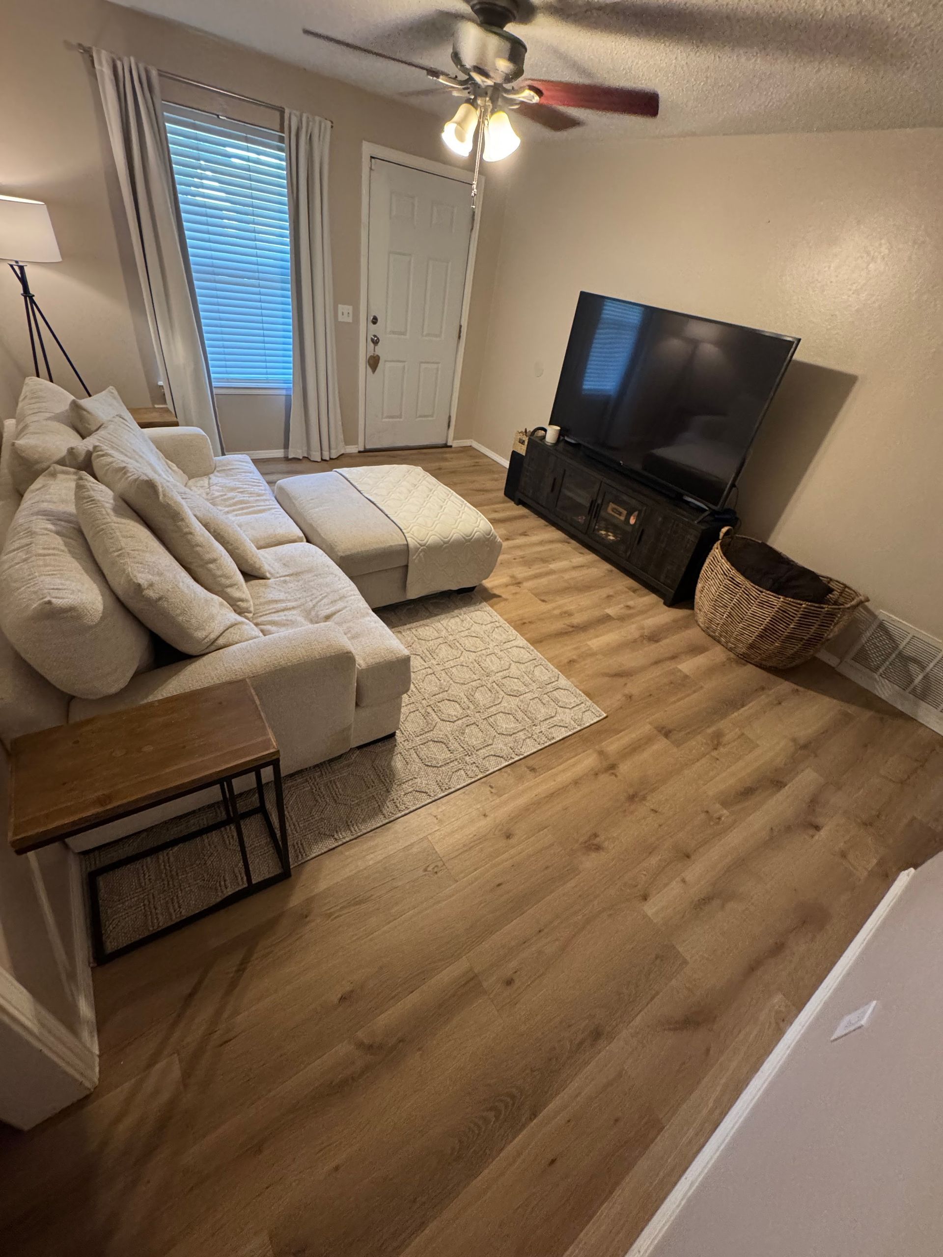 Bright living room with beige sectional, TV, white rug, wood floors, and ceiling fan near glass doors.