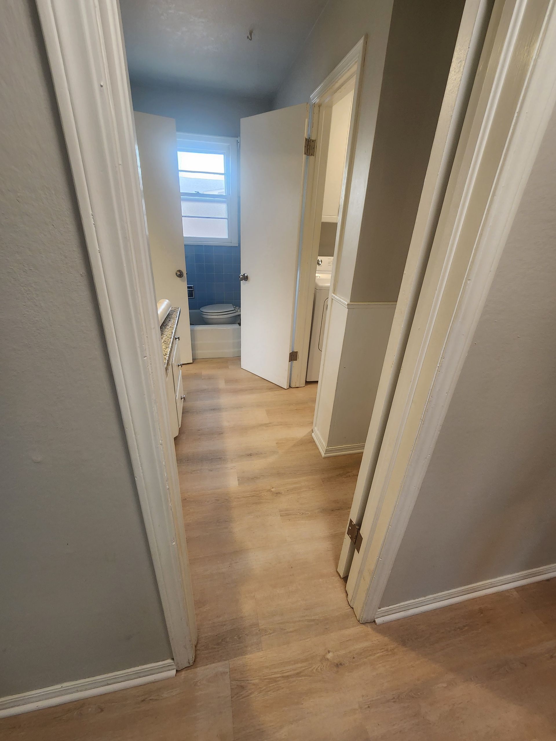Narrow hallway with light wood floors leading to an open bedroom door and window in the distance