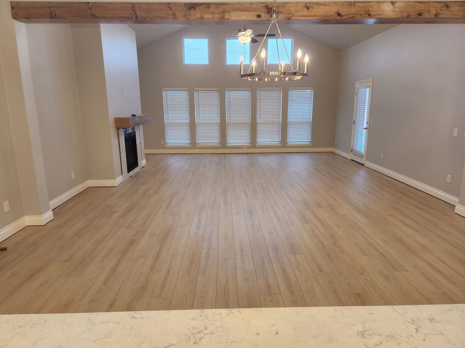 Empty living room with light wood floors, white walls, exposed beams, and chandelier.