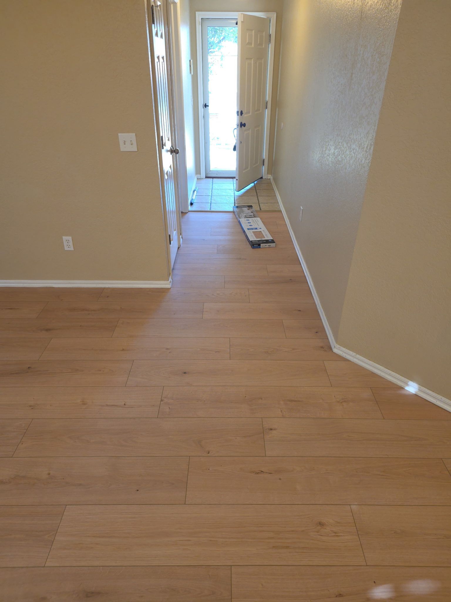 Empty hallway with light wood flooring and a doorway leading outside; construction materials lie near the door
