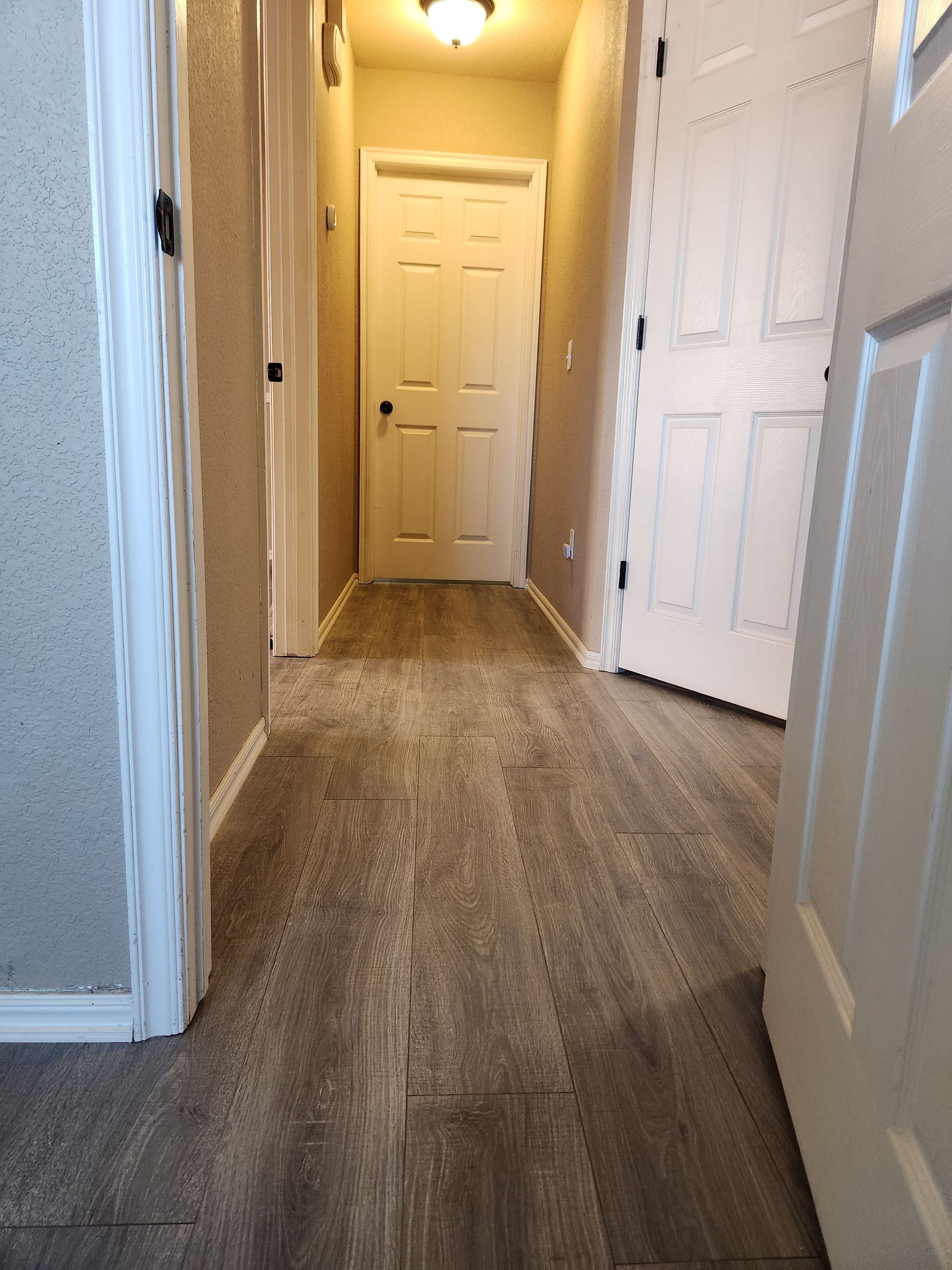 Narrow hallway with beige walls, wood-look flooring, and white doors at the end.
