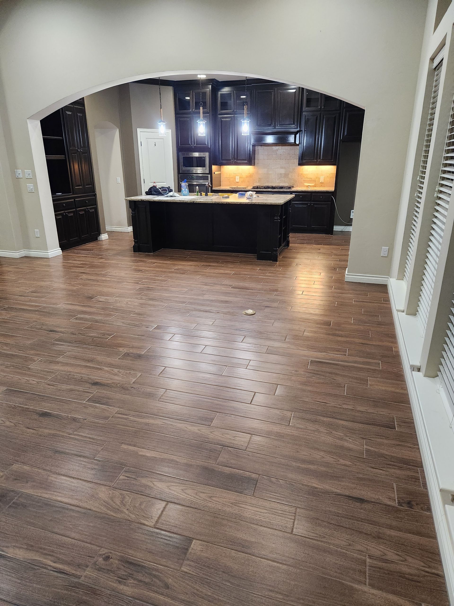 Empty room with wood floors and arched opening leading to a dark kitchen area with a central island.