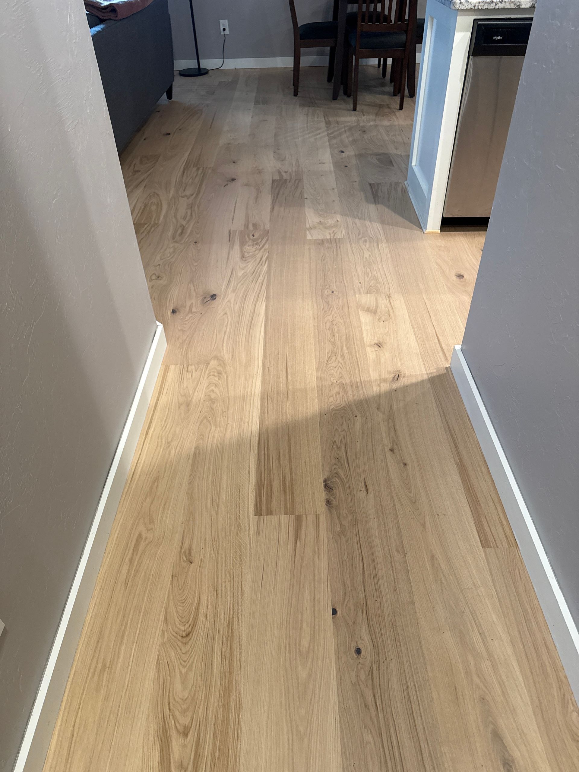 Narrow hallway with light wood floor leading into a room, with a small chair and furniture visible ahead