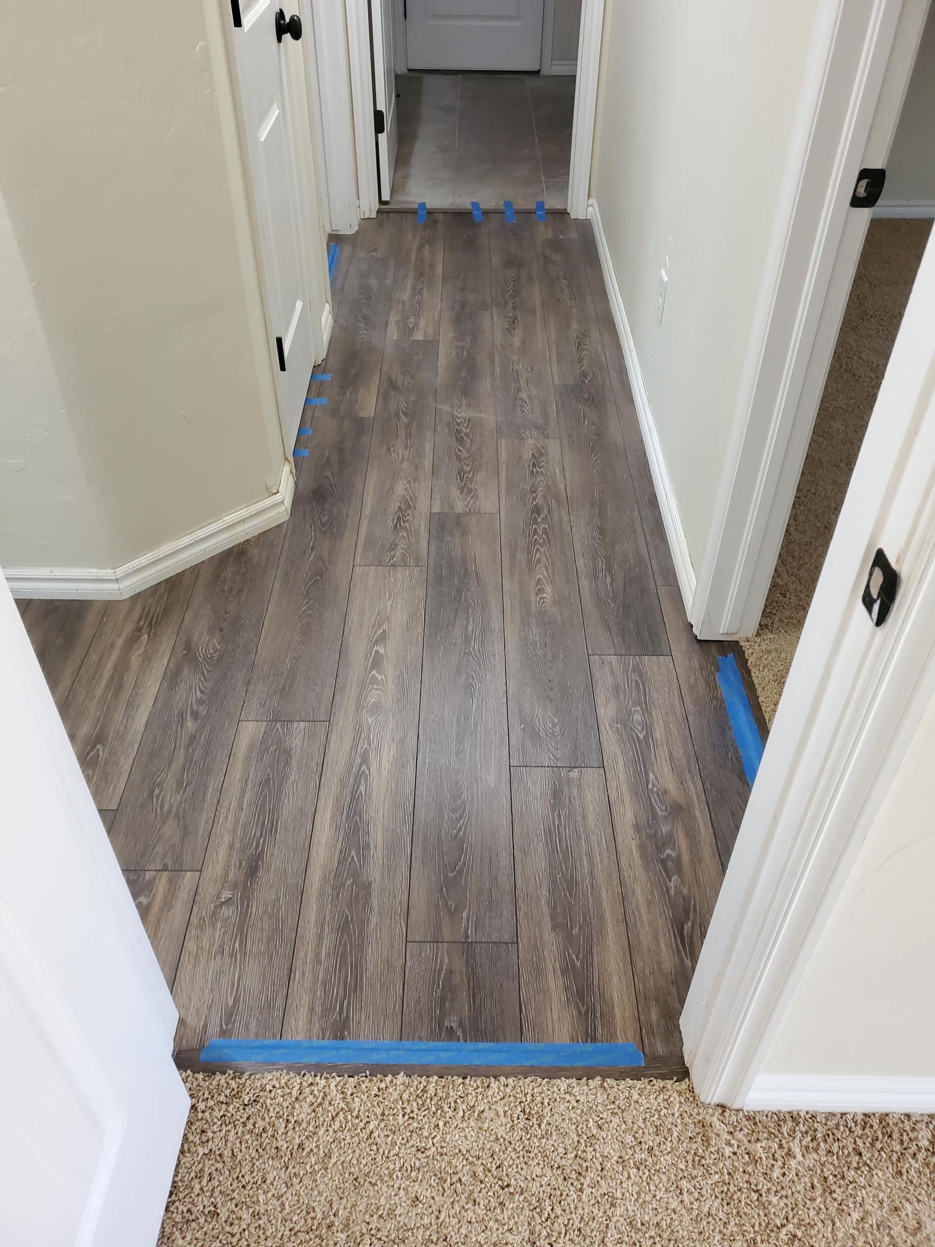 Narrow hallway with dark wood flooring, blue tape marking edges, and white walls and doors.