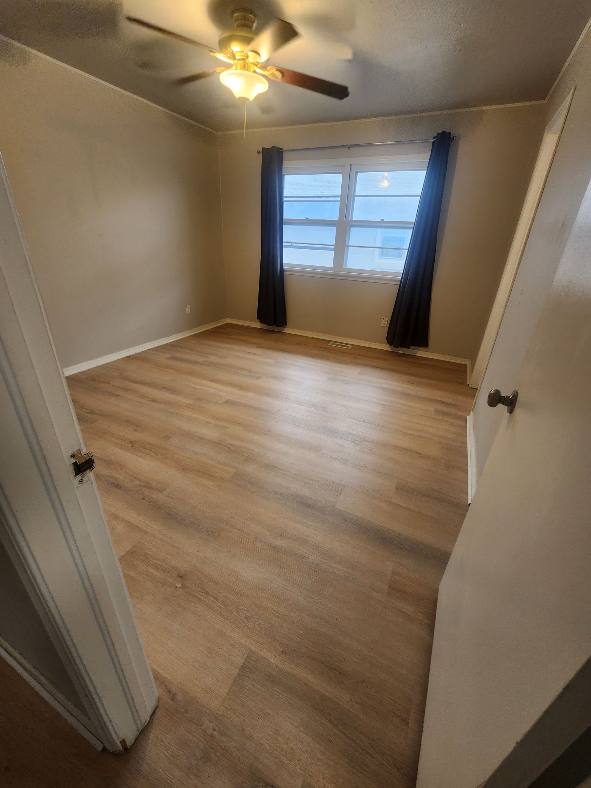 Empty beige bedroom with wood-look floor, ceiling fan, and window with dark curtains.