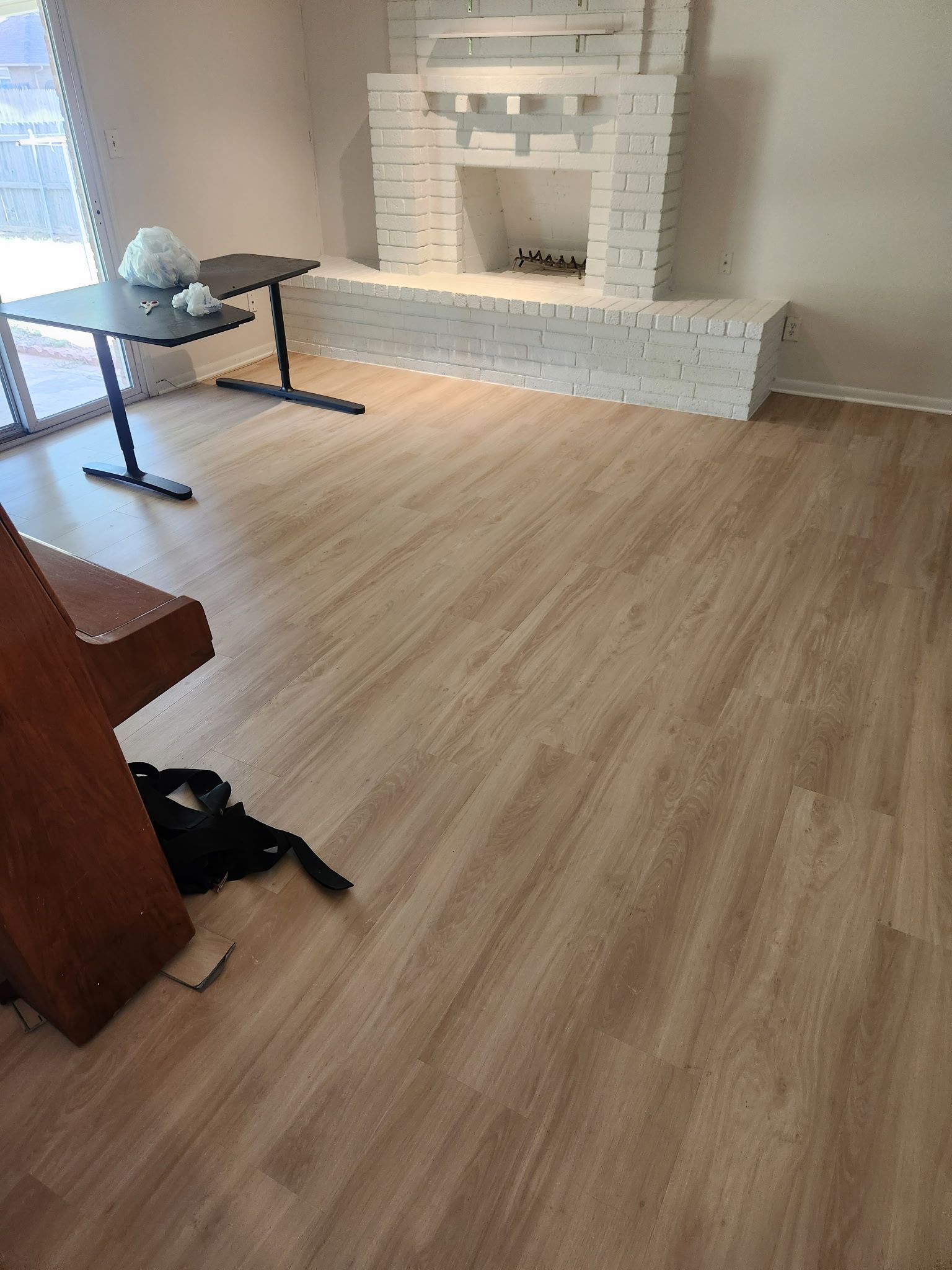 Empty room with light wood floor and a white brick fireplace, plus a chair and small table on the left.