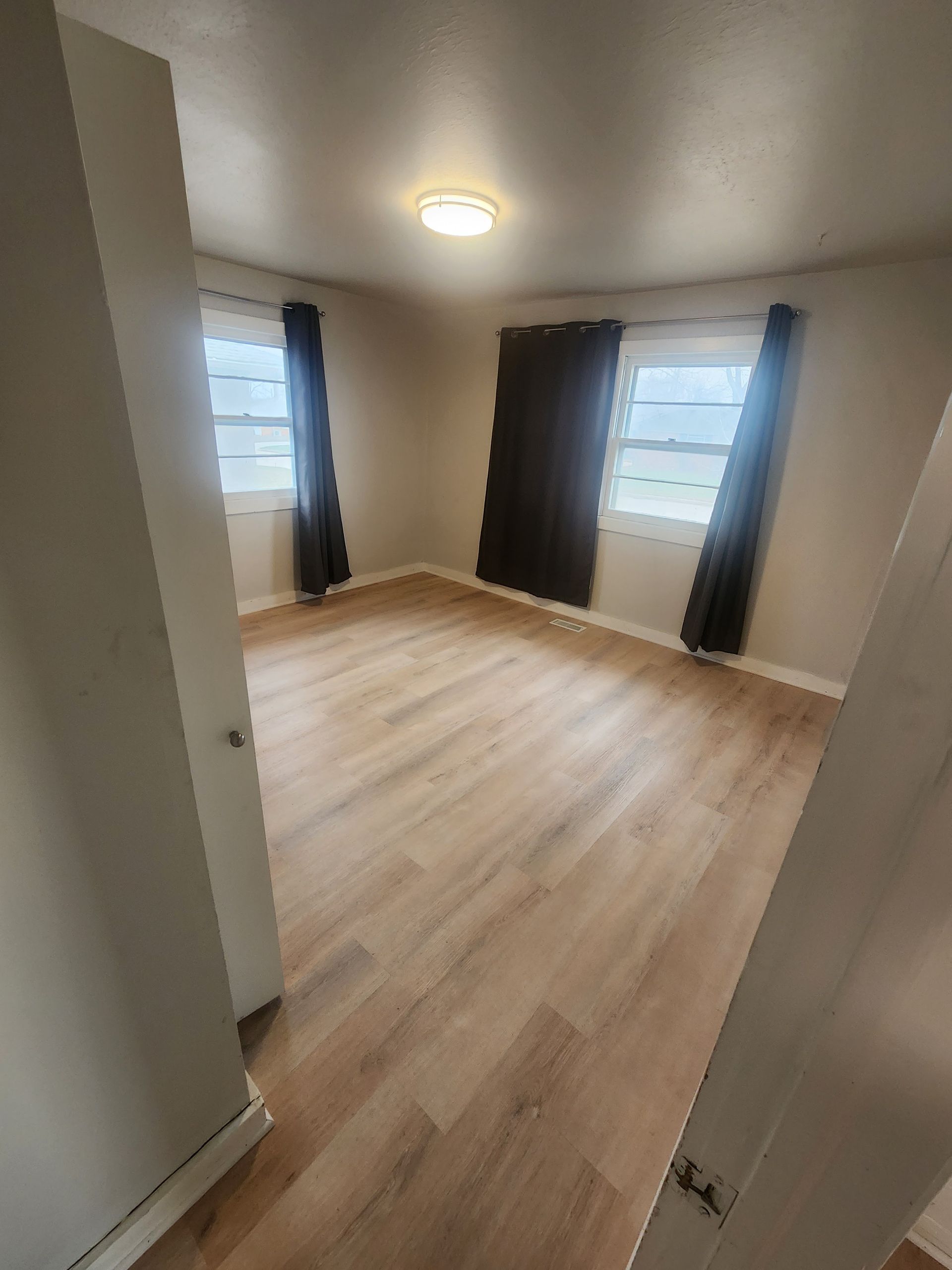 Empty bedroom with light wood floors, two windows, dark curtains, and a ceiling light.