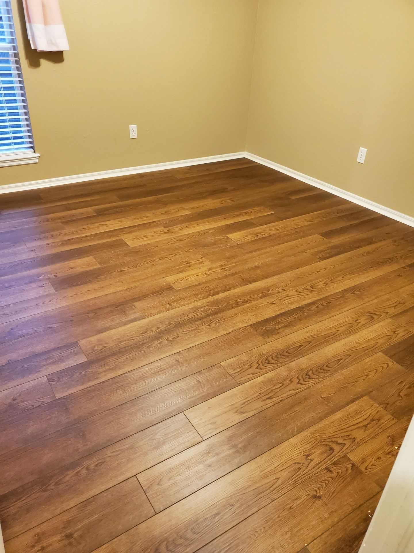 Empty room with wood floor, beige walls, and a window with blinds and curtain.