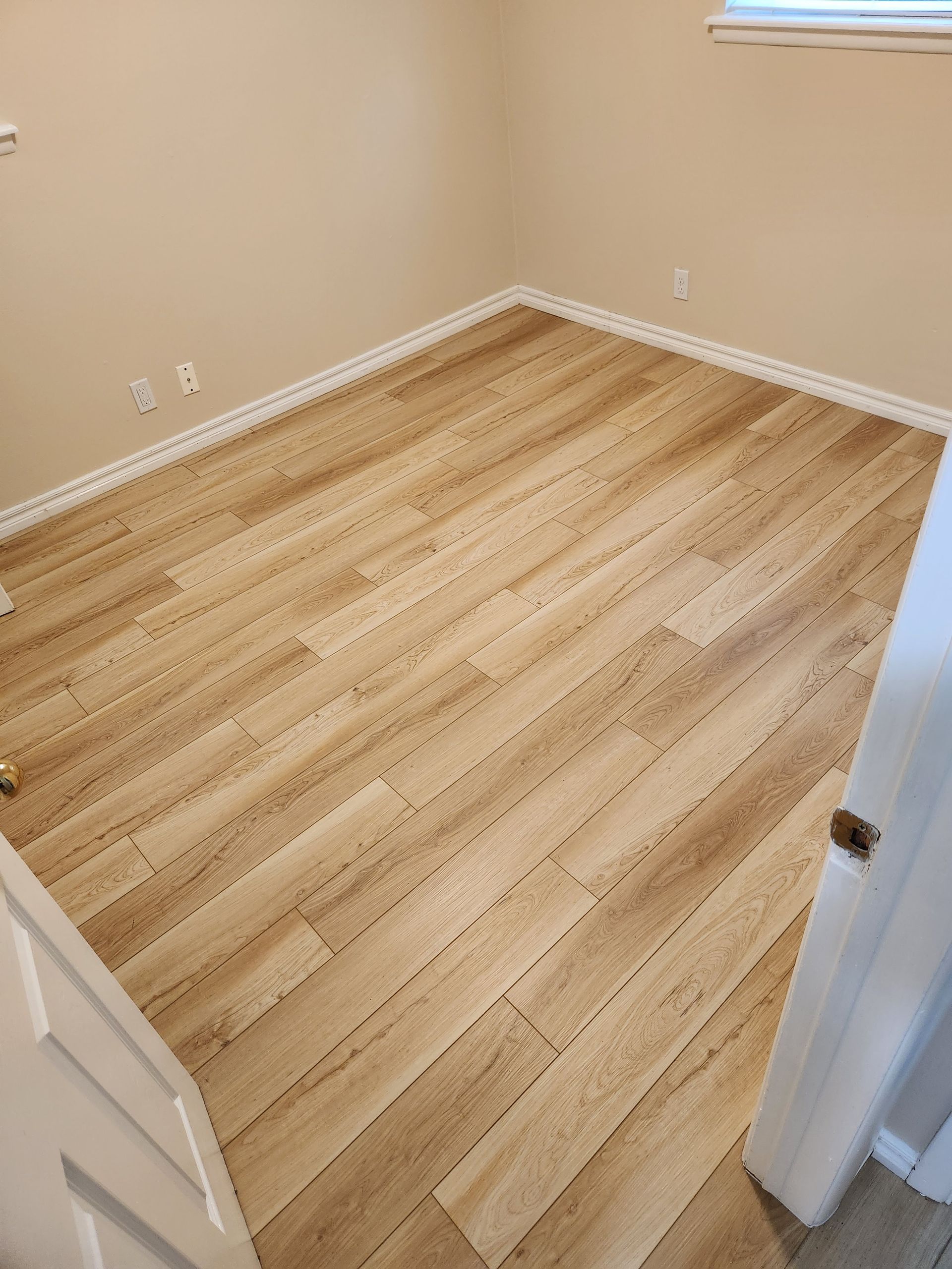 Empty beige room with light wood flooring and white trim, viewed through an open doorway