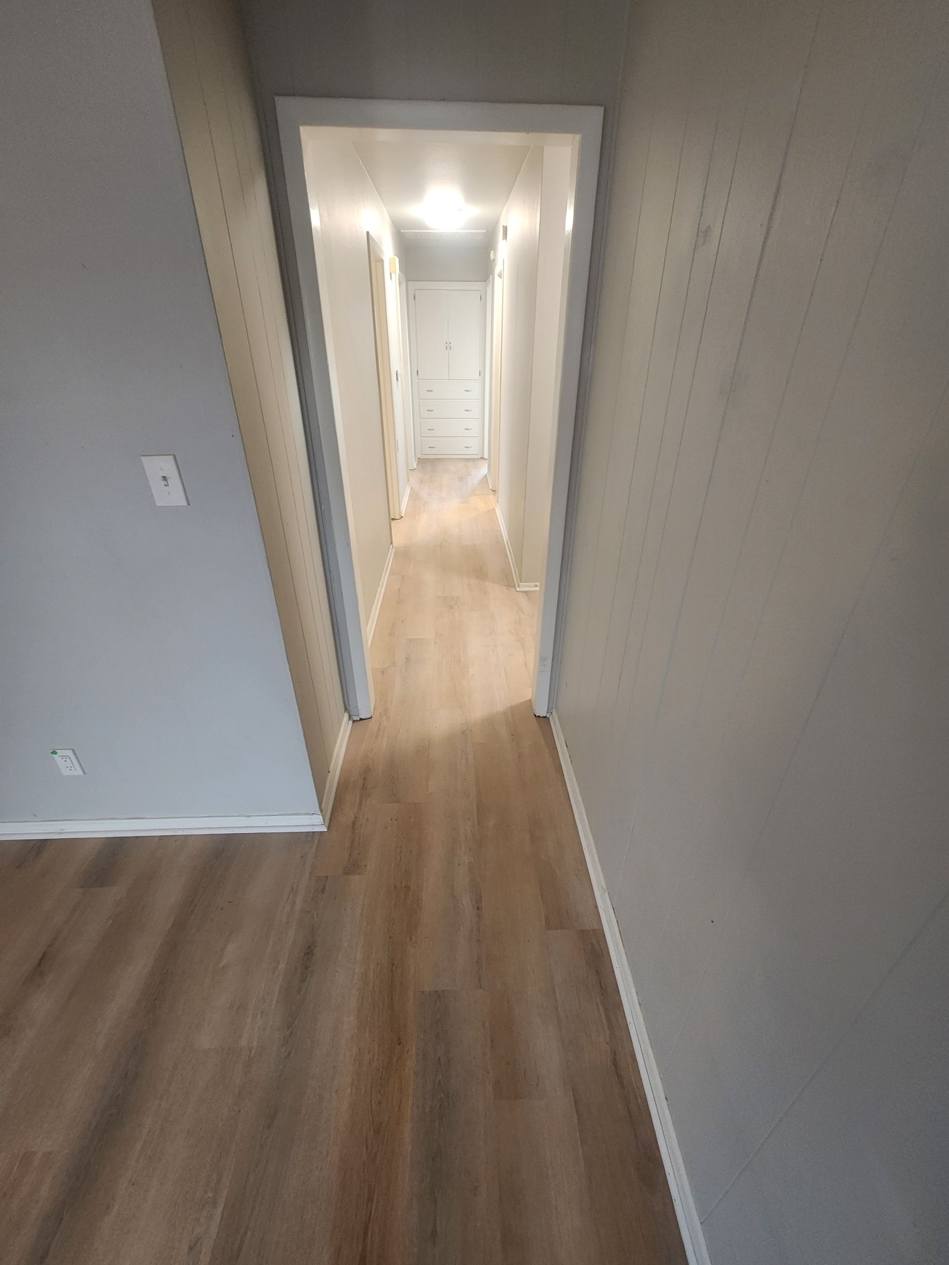Narrow gray hallway with wood flooring and white trim leading to a bright doorway.