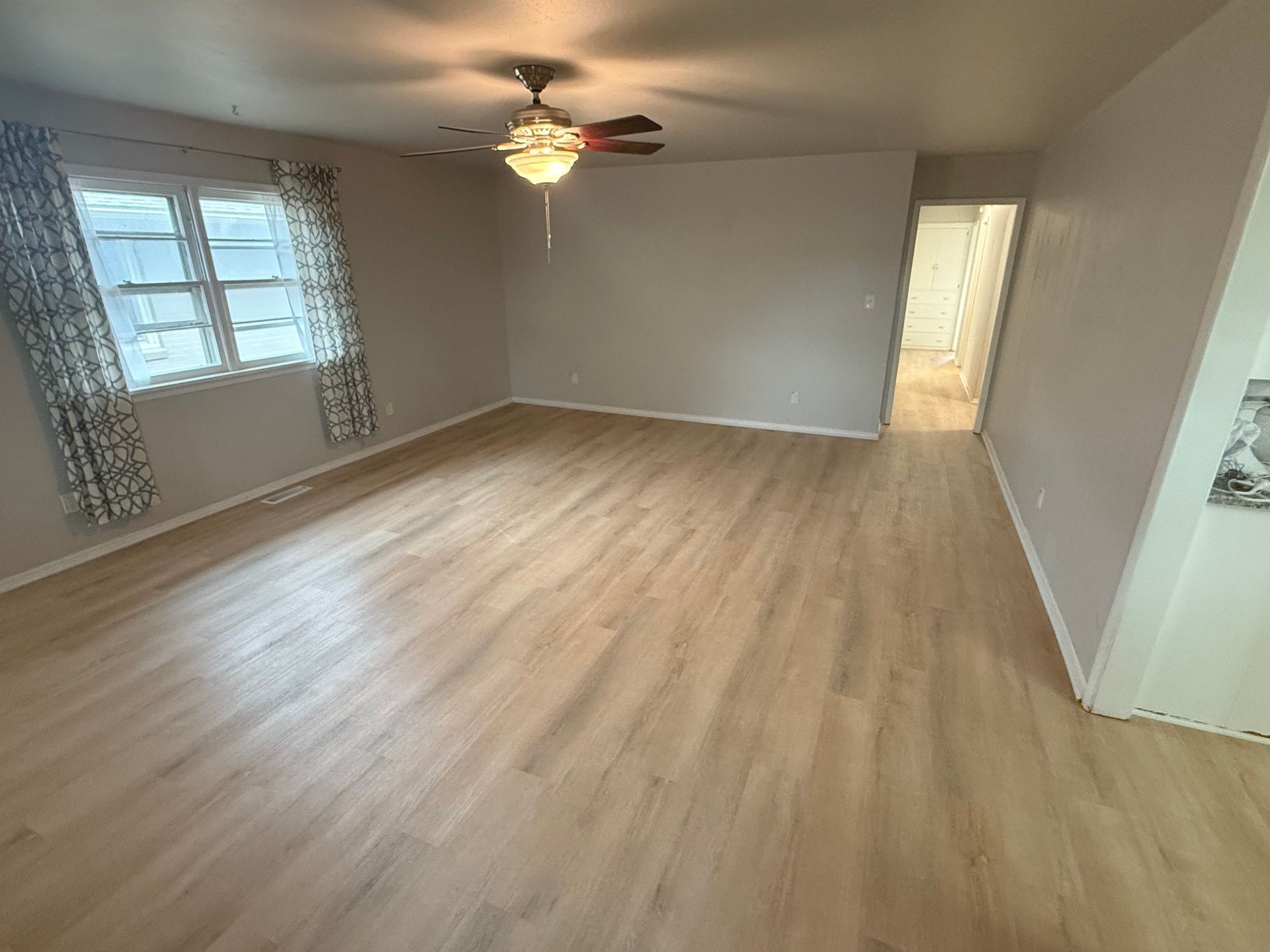 Empty living room with light wood floors, ceiling fan, gray walls, and windows with sheer curtains.