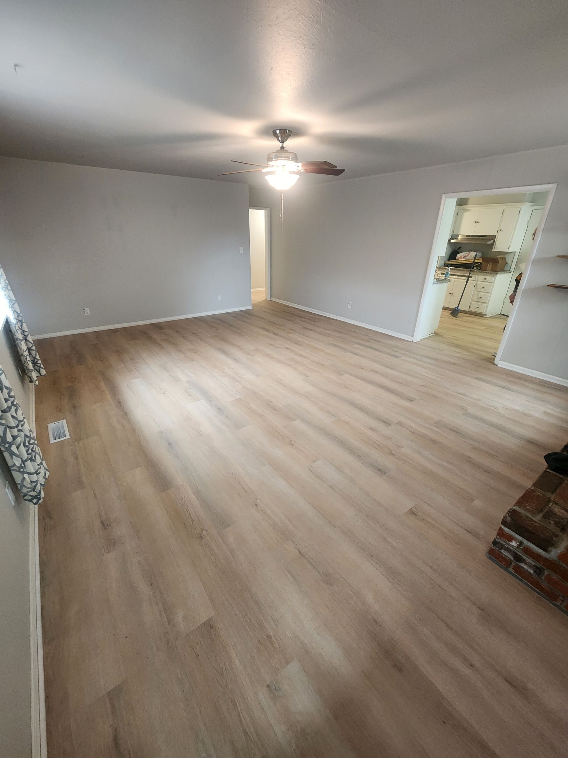 Empty living room with gray walls, light wood floors, and a ceiling fan; kitchen visible through doorway.