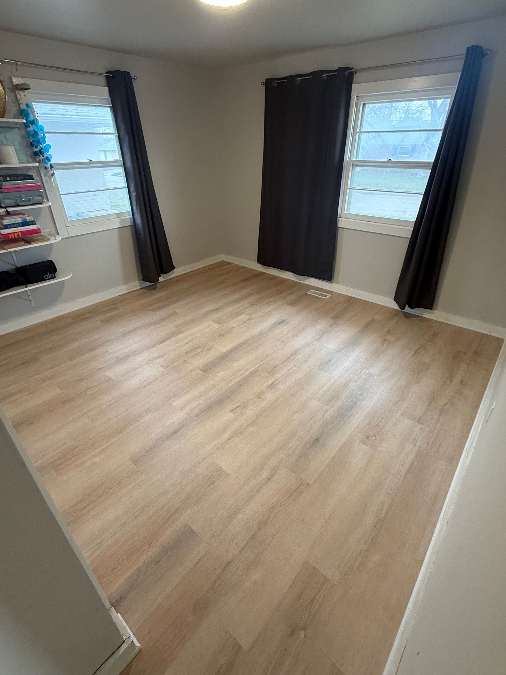 Empty bedroom with light wood floor, white walls, and two windows with dark curtains.