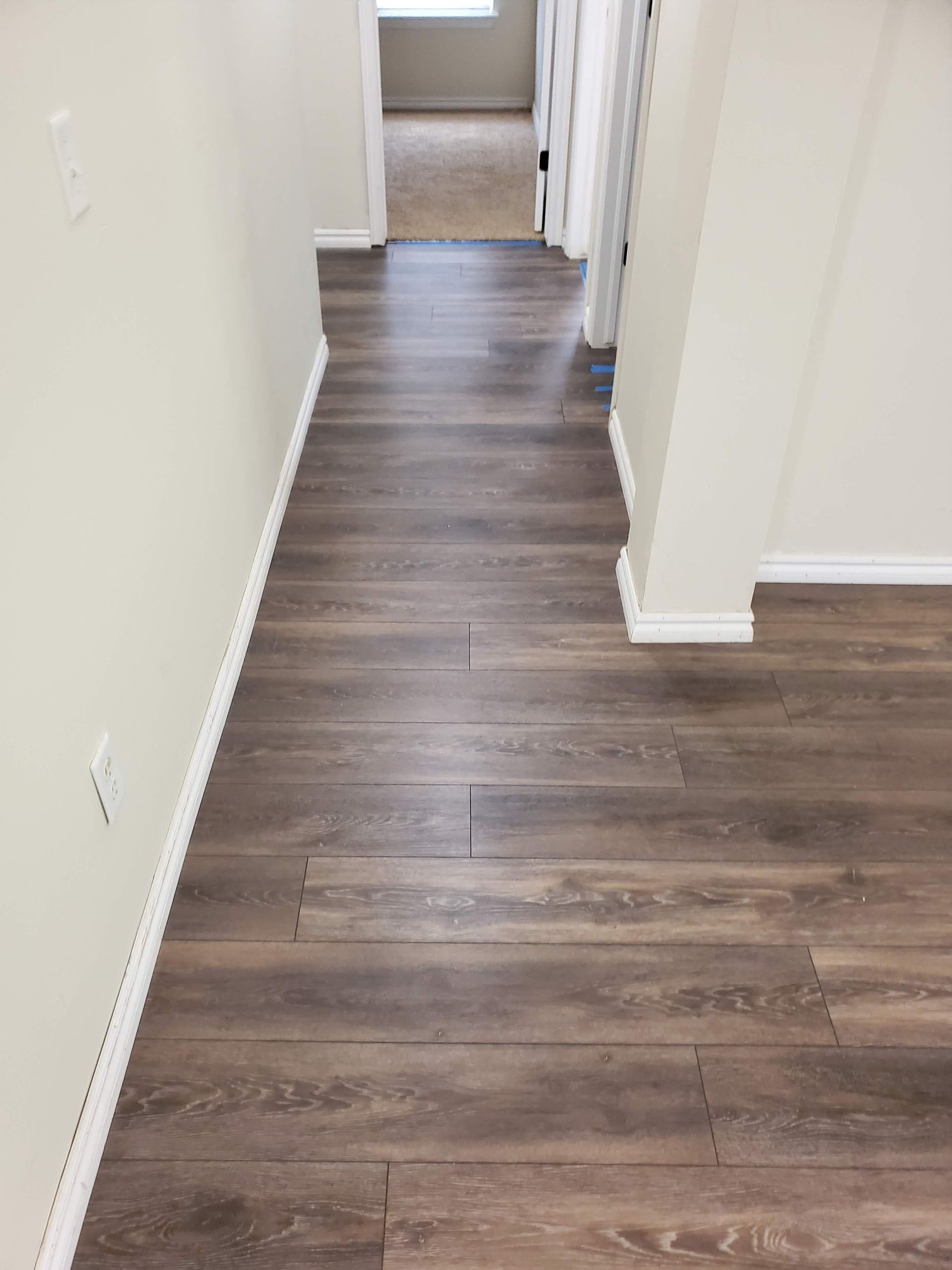 Empty hallway with brown wood flooring and white walls leading to a carpeted room