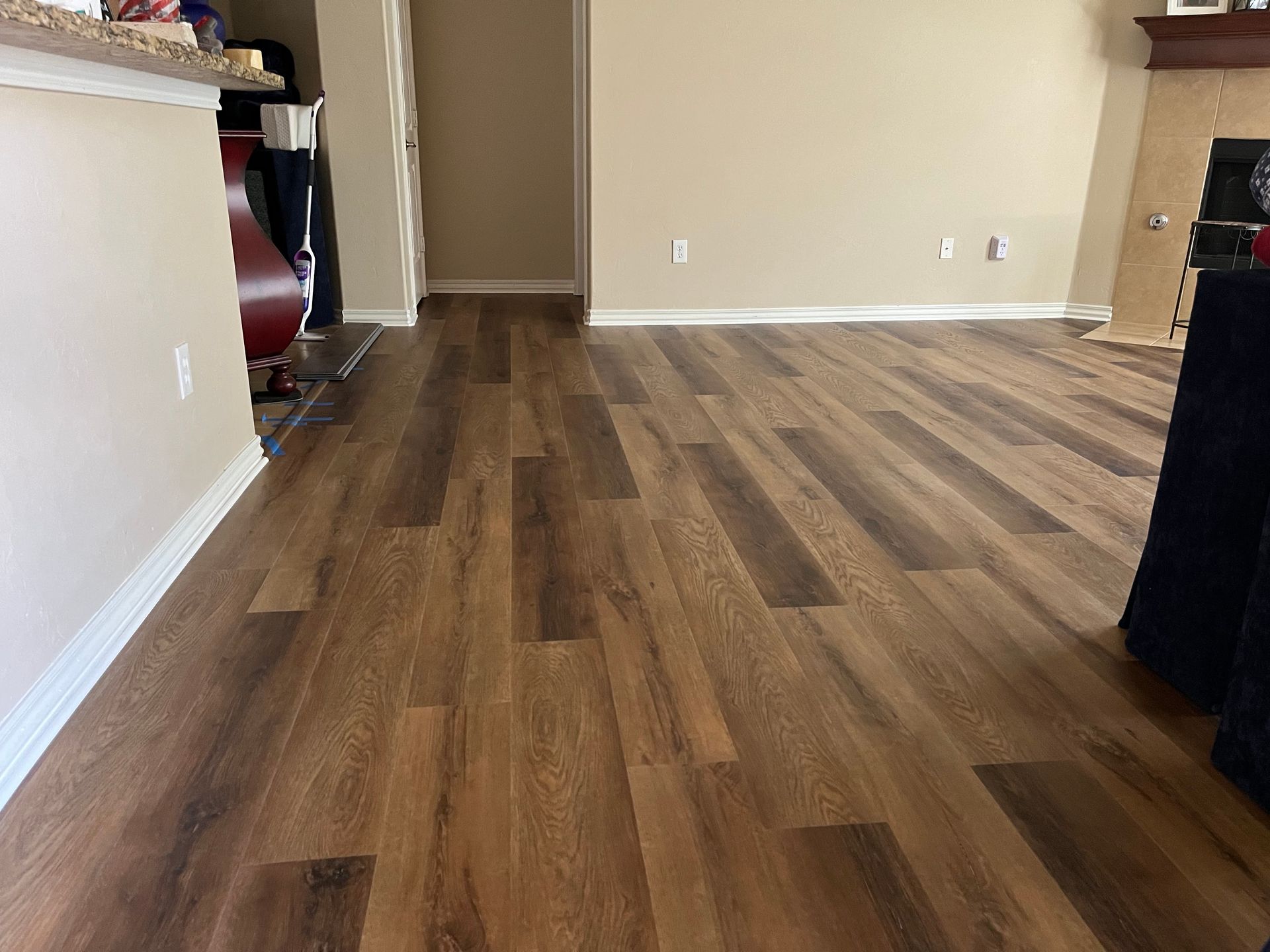 Empty living room with wood plank flooring and beige walls, viewed toward a hallway and fireplace.