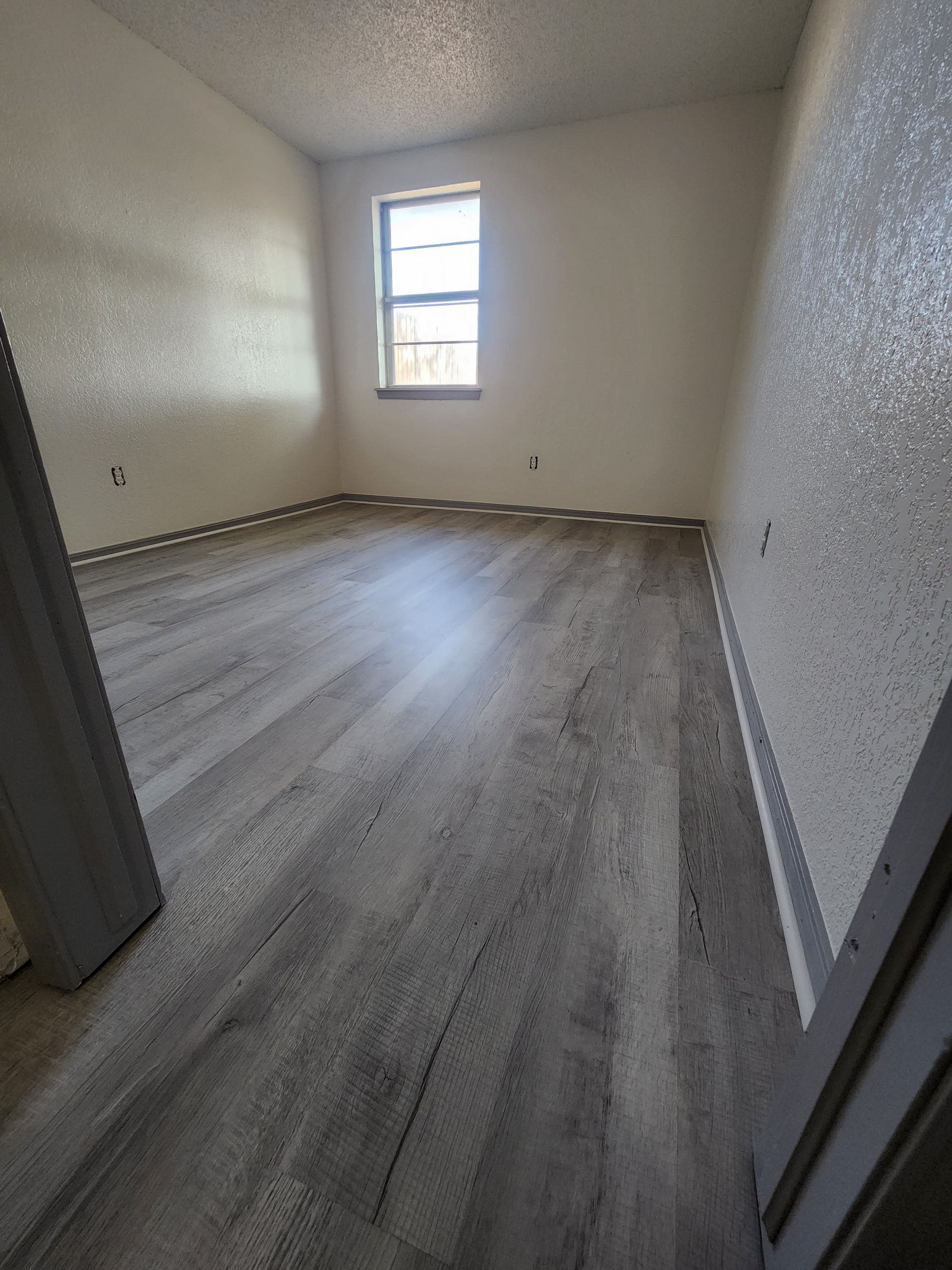 Empty small room with gray wood flooring, white textured walls, and one narrow window.