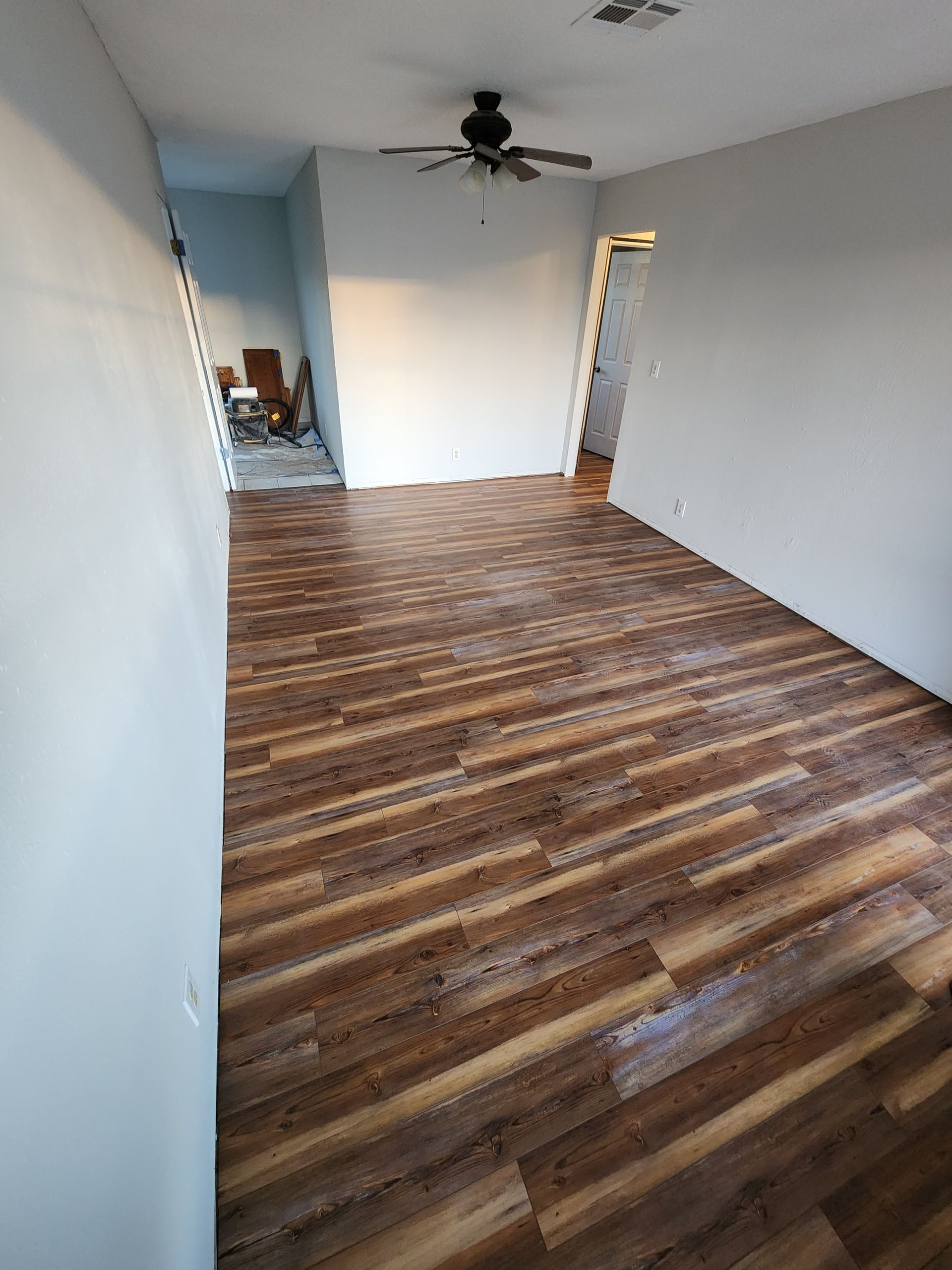 Empty room with wood-look flooring, white walls, and a ceiling fan near an open doorway.