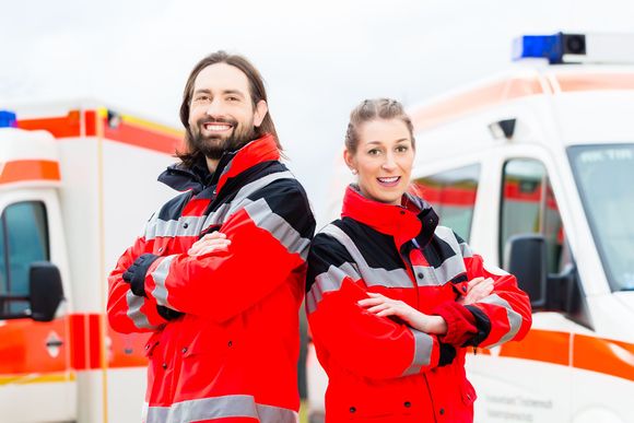 Two emergency medical technicians smiling in front of ambulances, arms crossed.