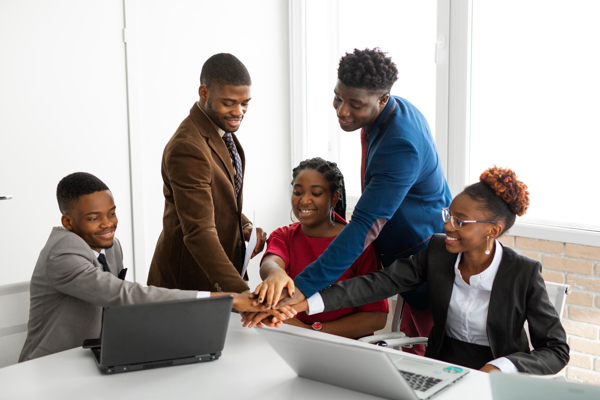 Five people in business attire with hands stacked, celebrating a team achievement, indoors near laptops.