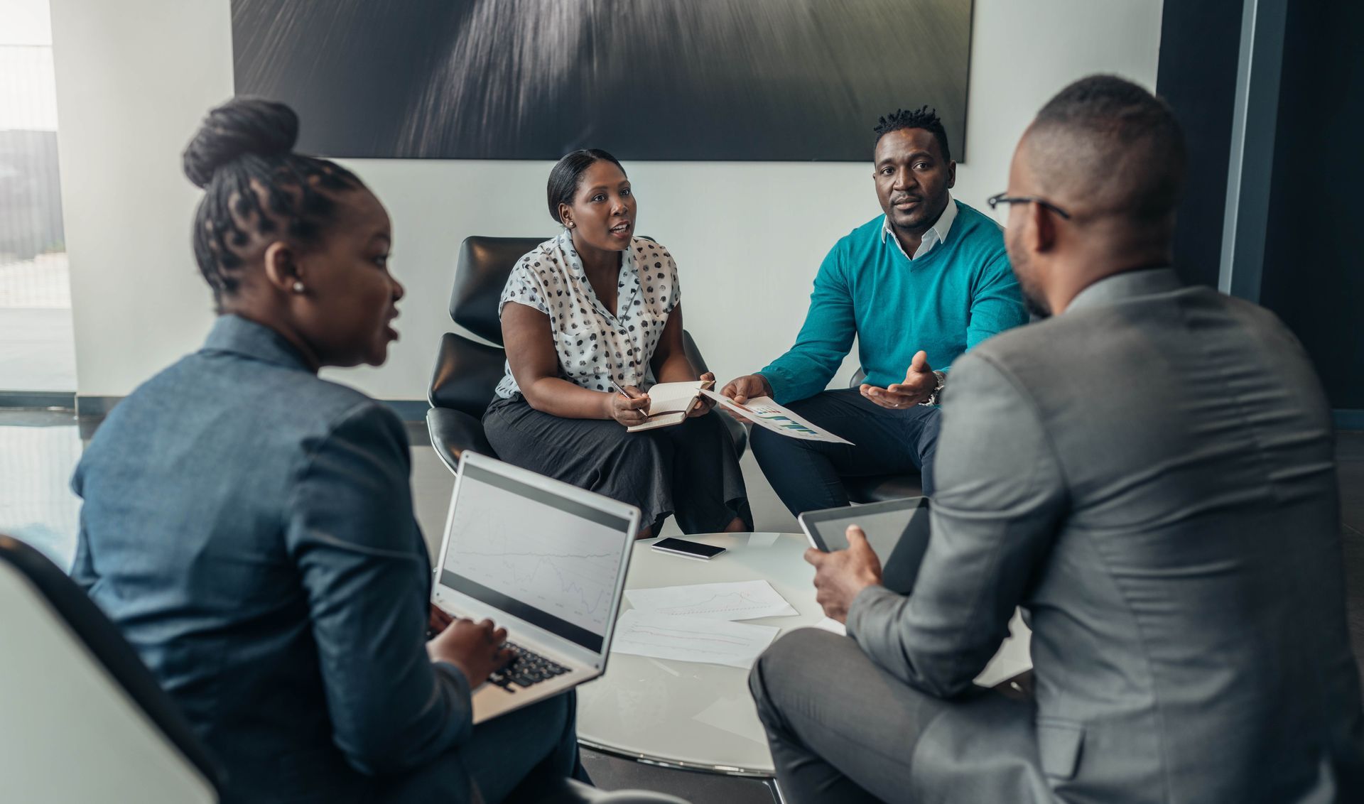 Four businesspeople in a circle, discussing documents. One uses a laptop, others have papers or a tablet. Modern office setting.