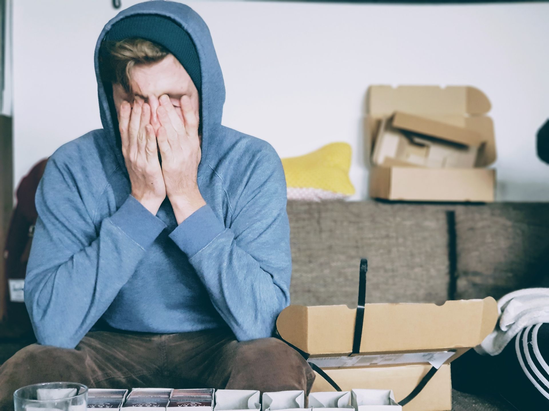 Person in blue hoodie covering face, looking stressed, sitting on a couch. Boxes in background.