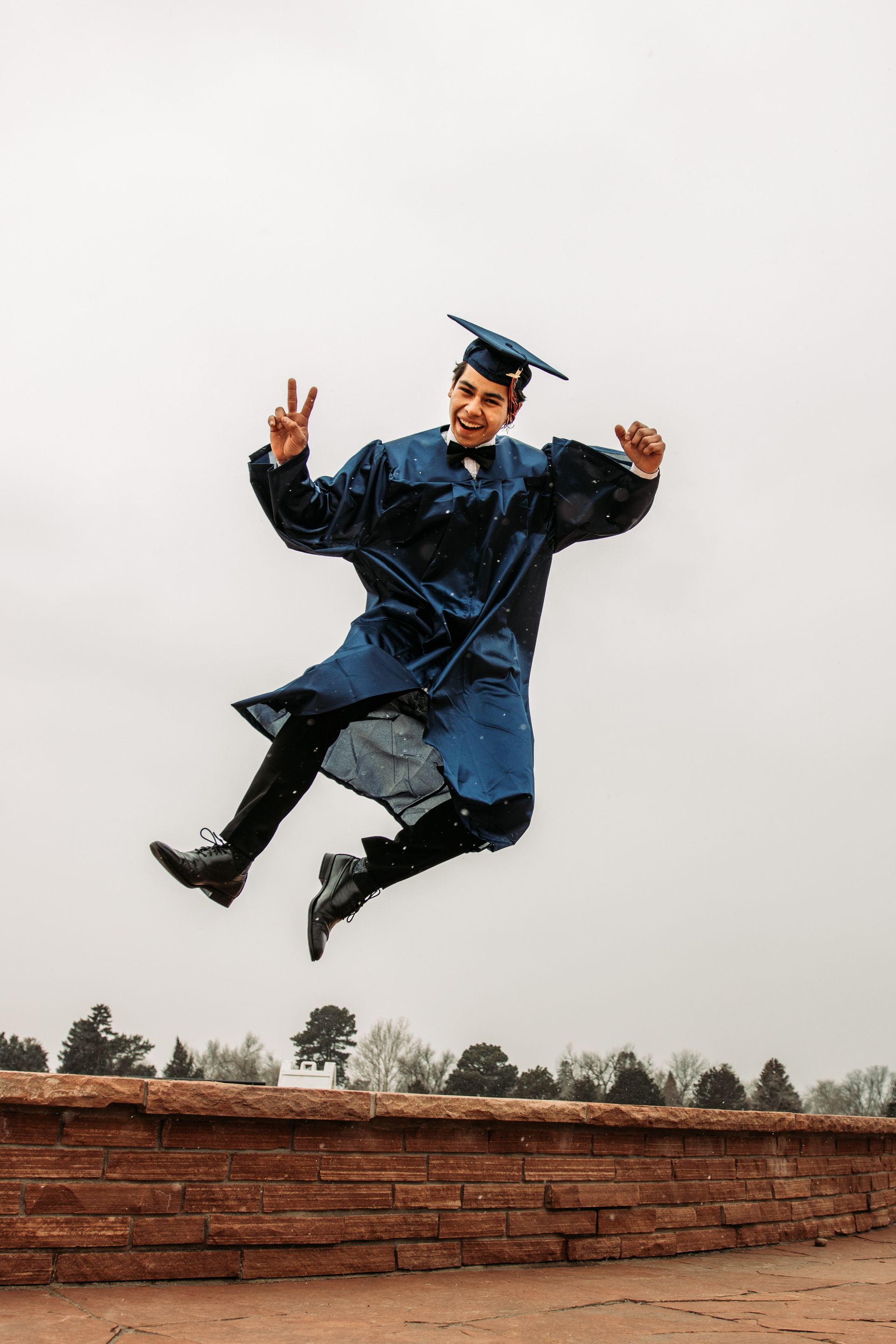 Person in blue graduation gown jumps with joy, making peace sign.
