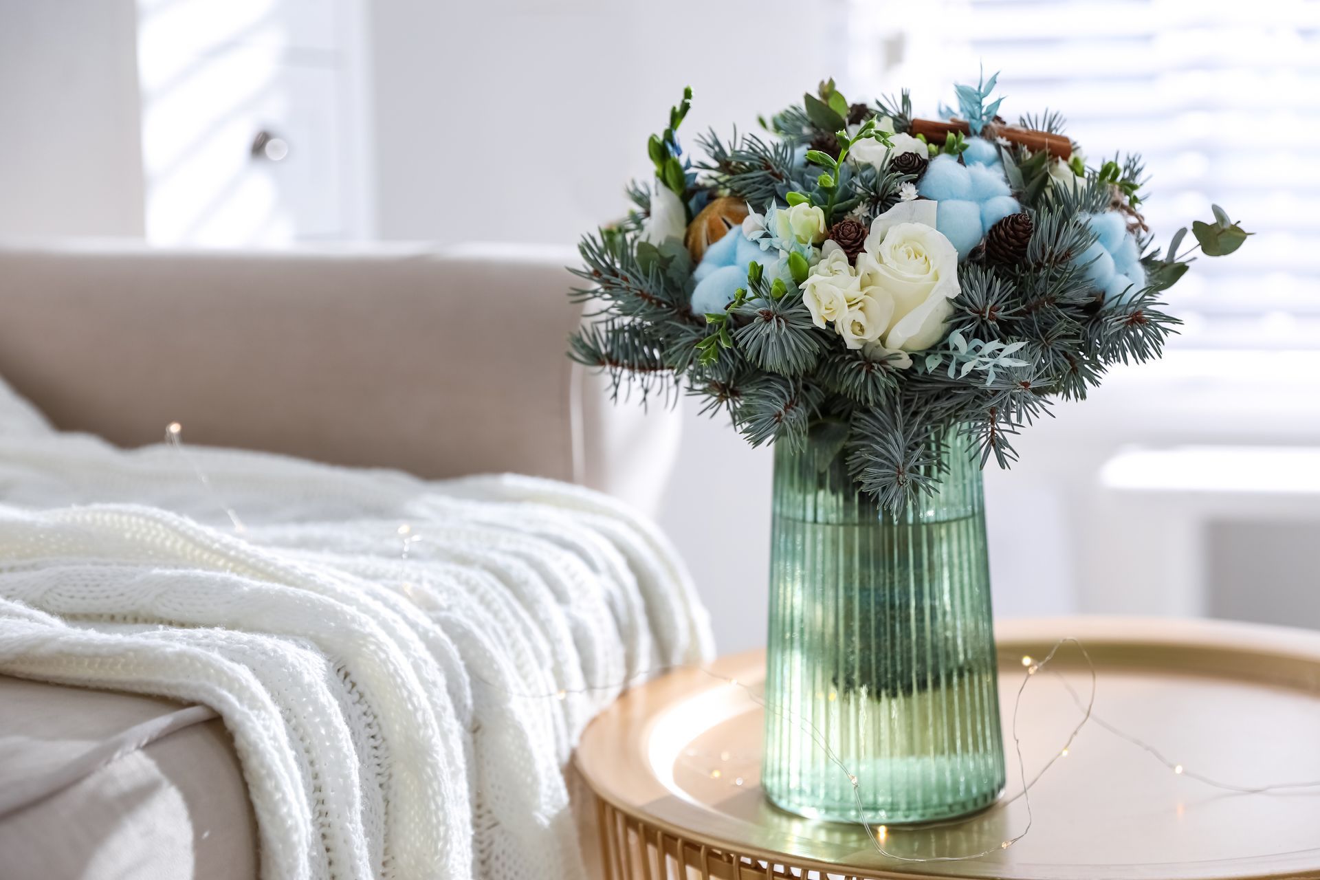 Bouquet of flowers in a green vase on a gold table with a cozy blanket and couch in the background.