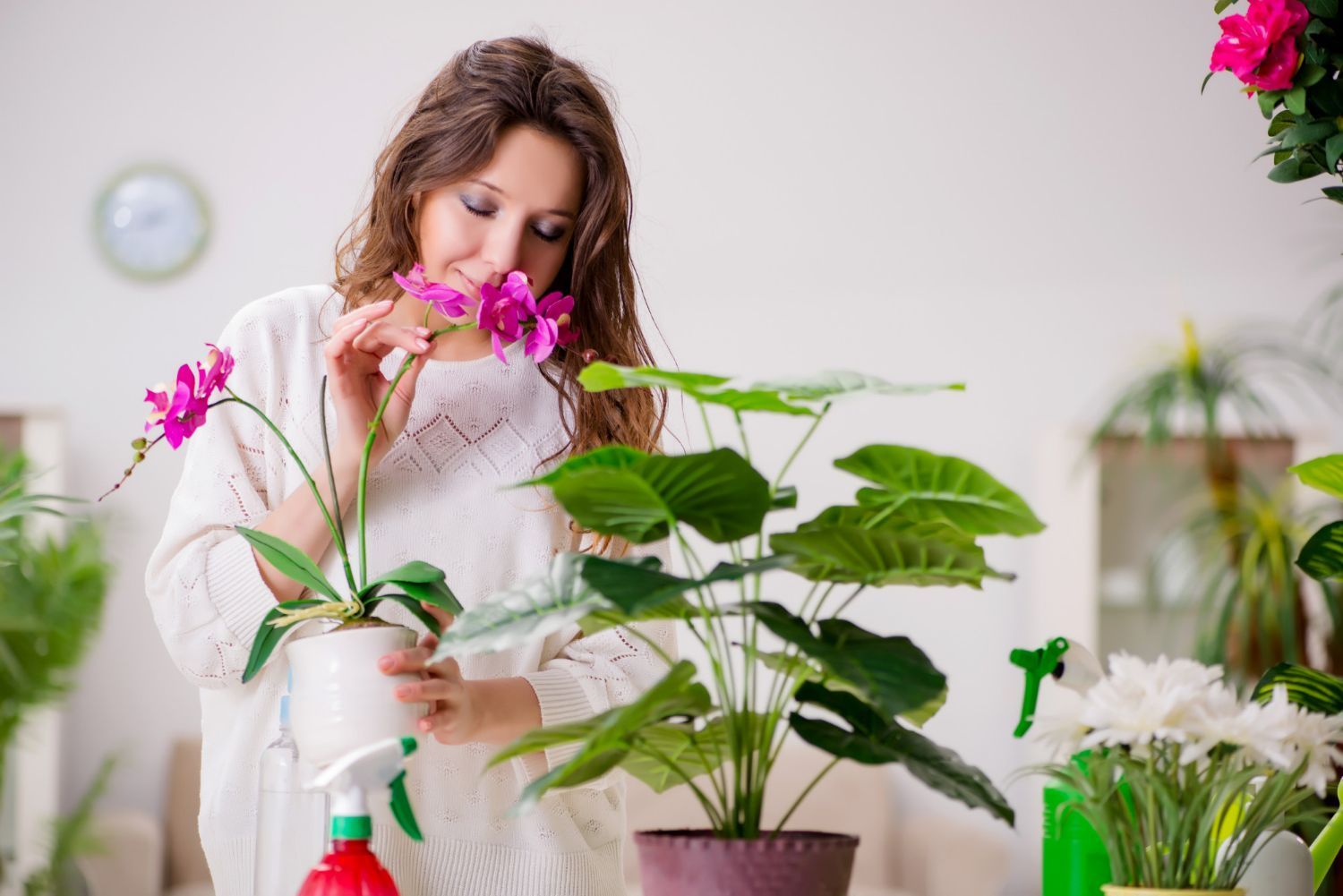 A woman is smelling a flower while watering a potted plant.