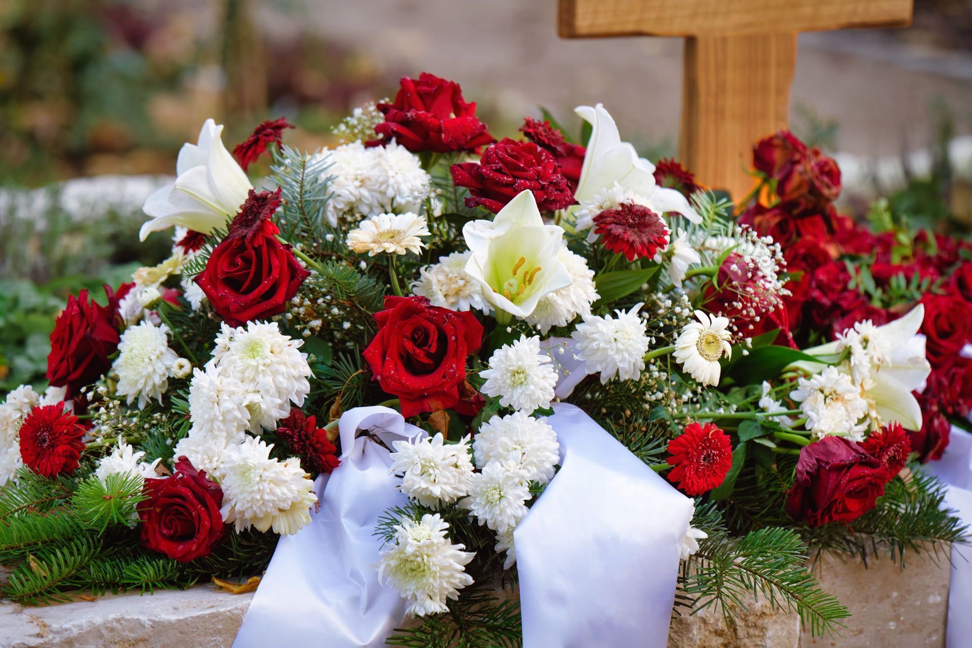 Red and white flowers, including roses and lilies, on a grave, with a cross in the background.