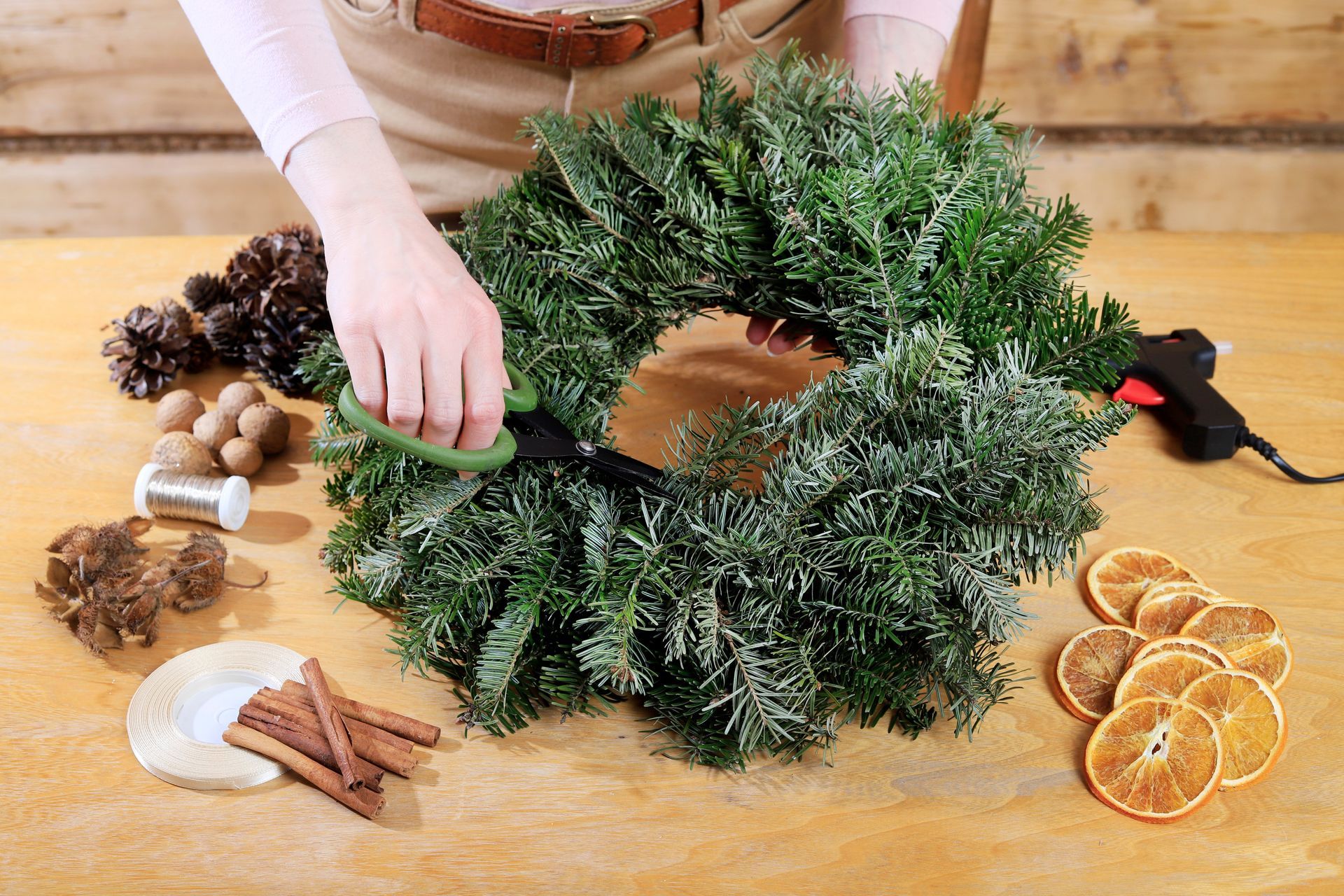 Person crafting a Christmas wreath with greenery, orange slices, and pine cones on a wooden table.