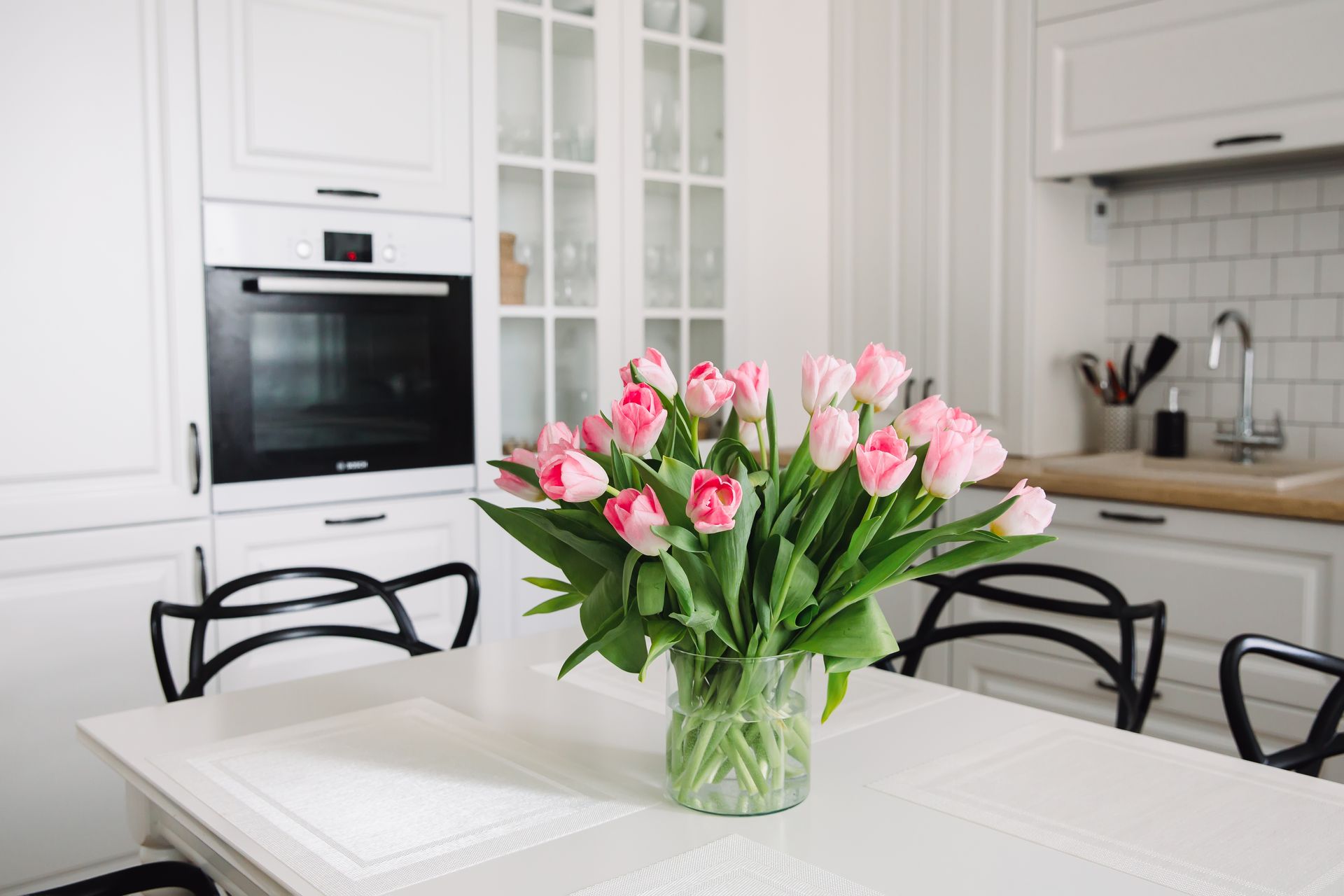 Pink tulips in a glass vase on a white table in a bright kitchen with white cabinets and black chairs.