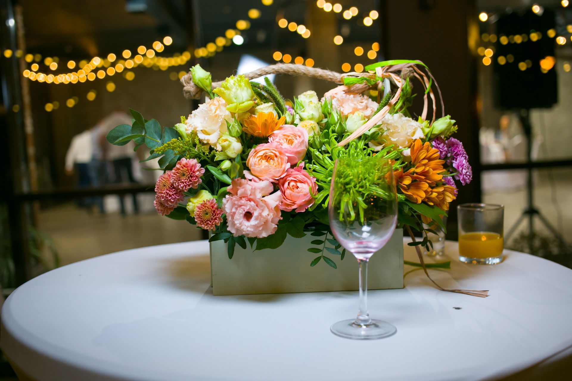 Floral arrangement in a wooden box on a table, with a wine glass, and lights in the background.