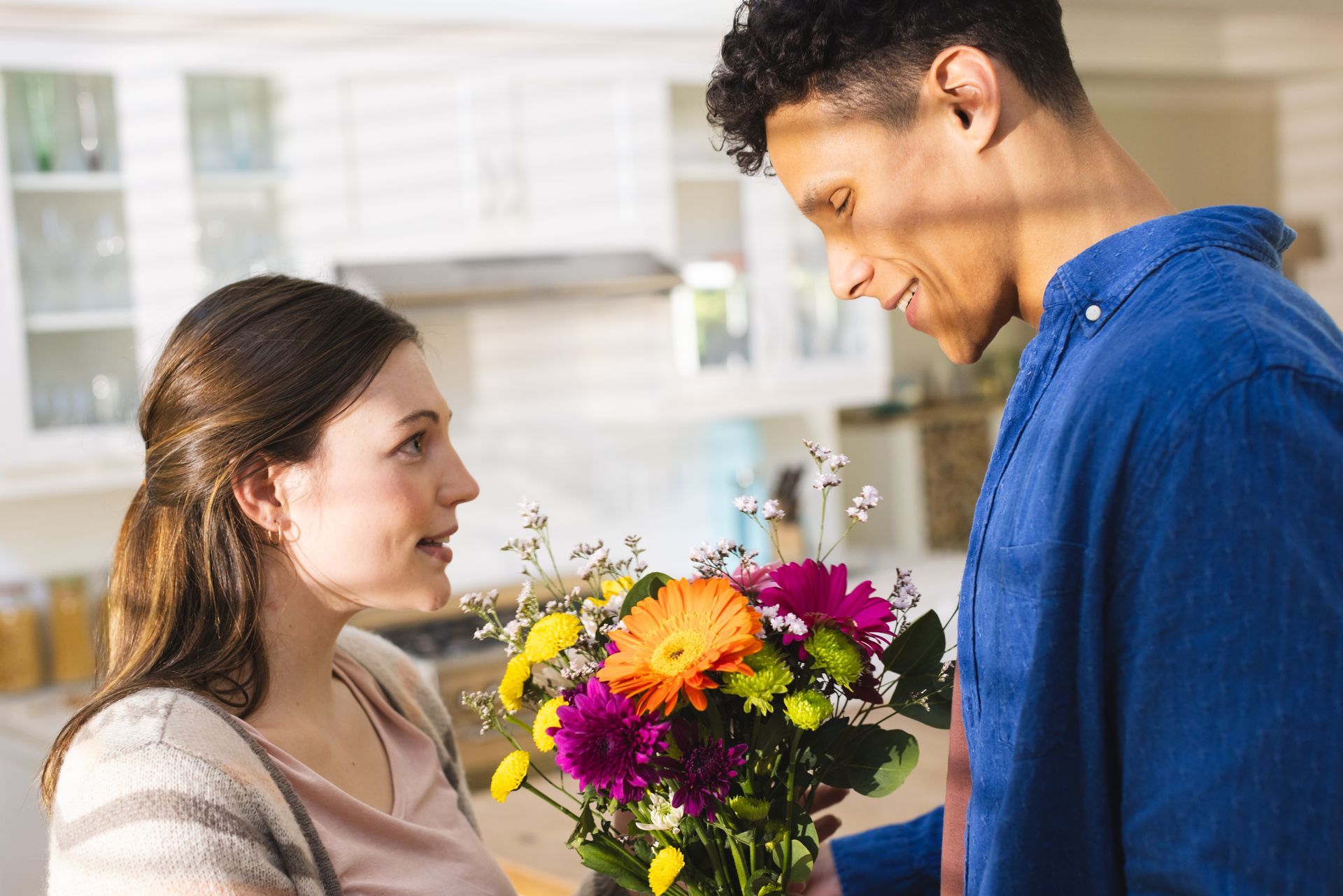 Man giving woman colorful flowers in a kitchen, both smiling.