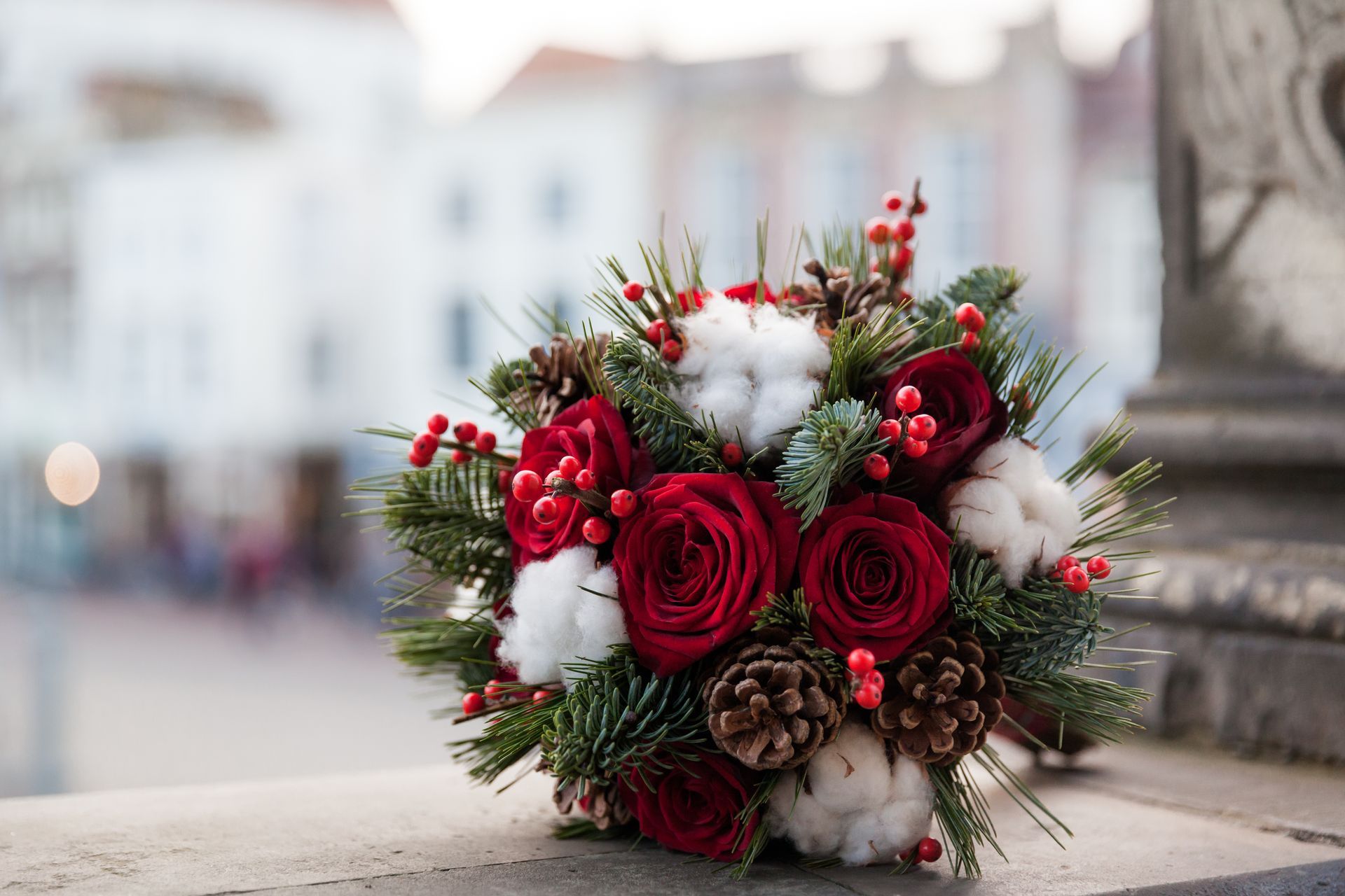 A bouquet of red roses , cotton , pine cones and berries is sitting on a stone wall.