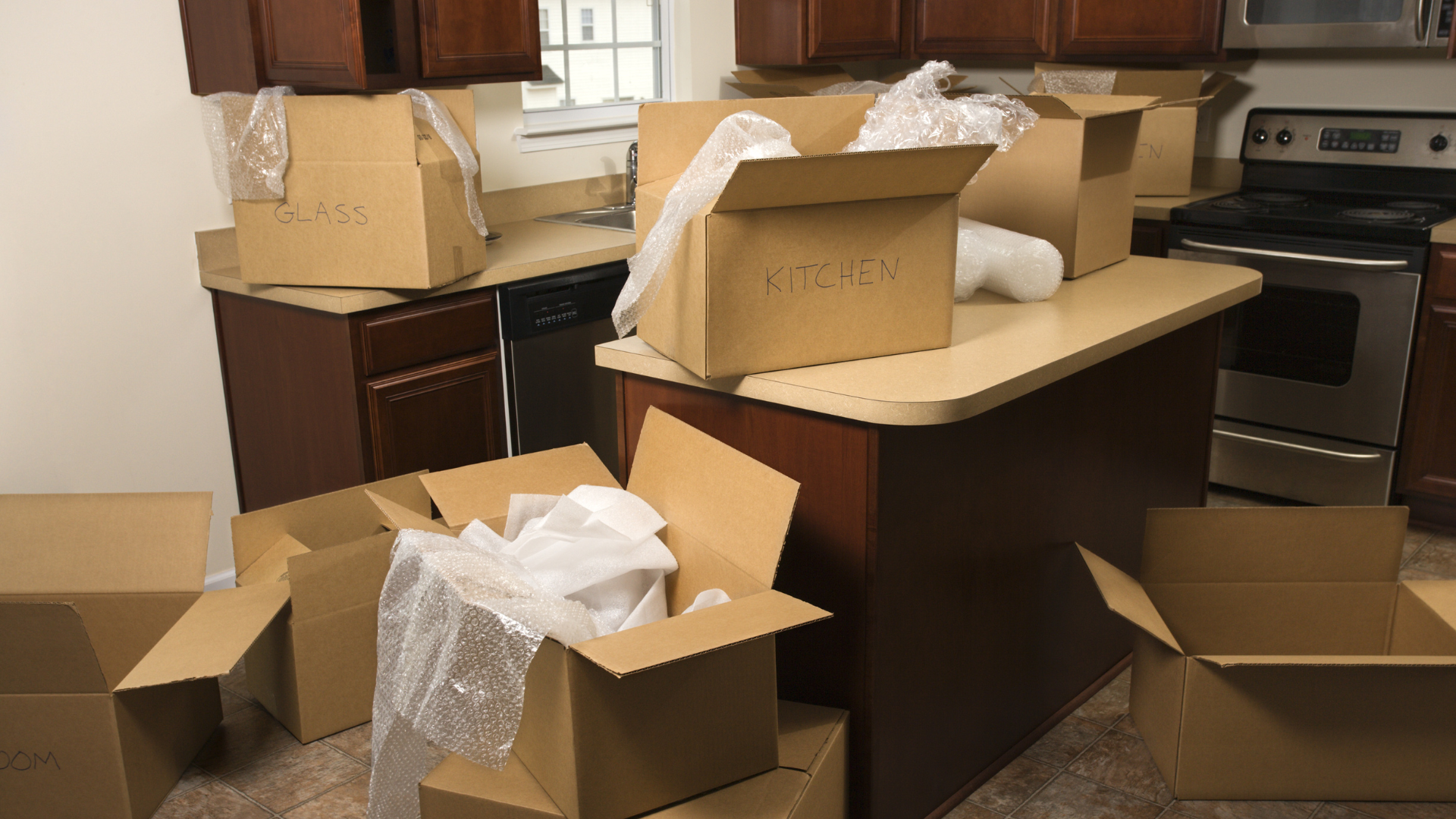Kitchen with packed cardboard boxes, preparing for a move. Brown cabinets and countertops, appliances in background.