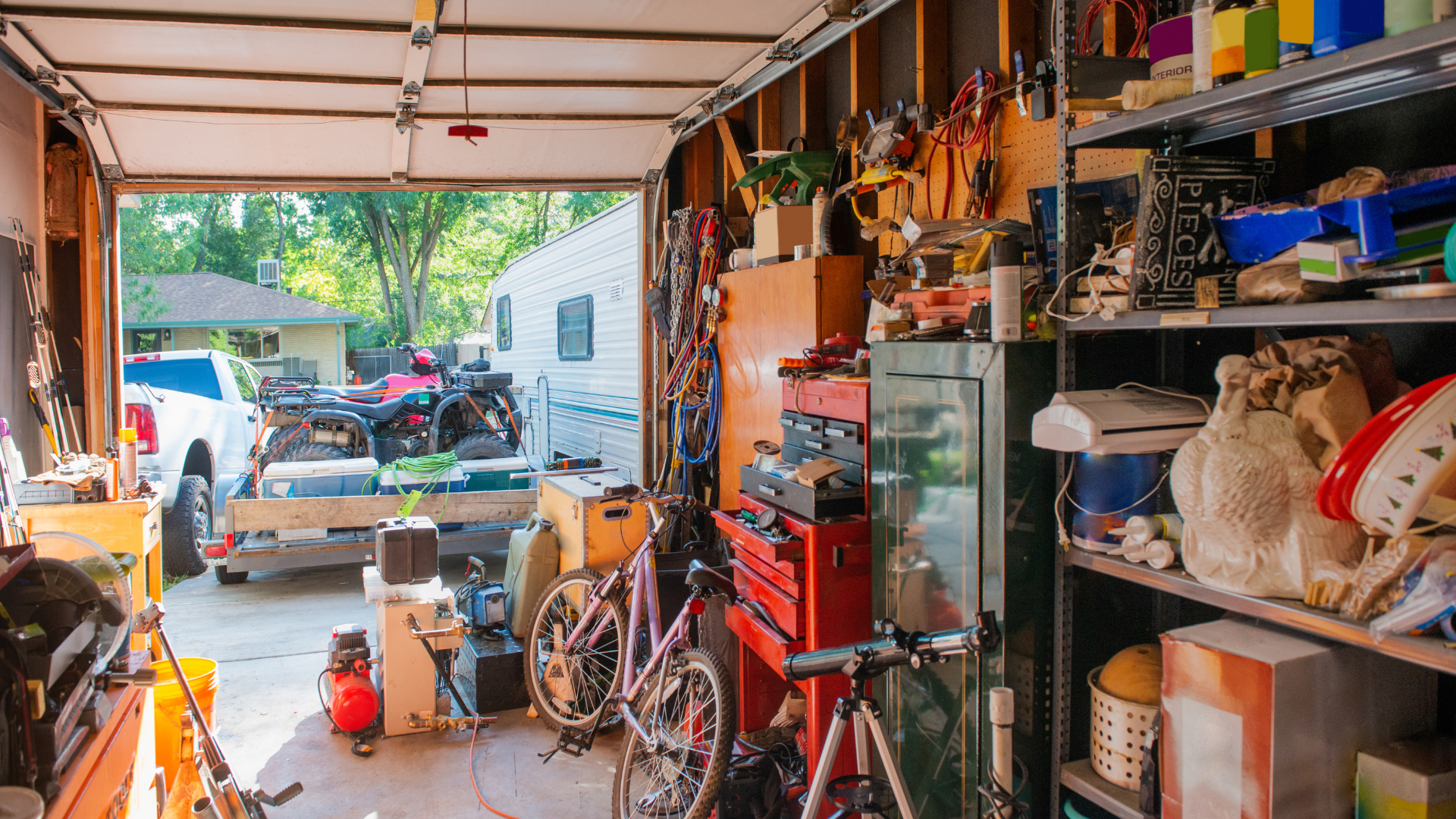 Cluttered garage with open door; bicycles, tools, and shelves of items. View of vehicles and houses outside.