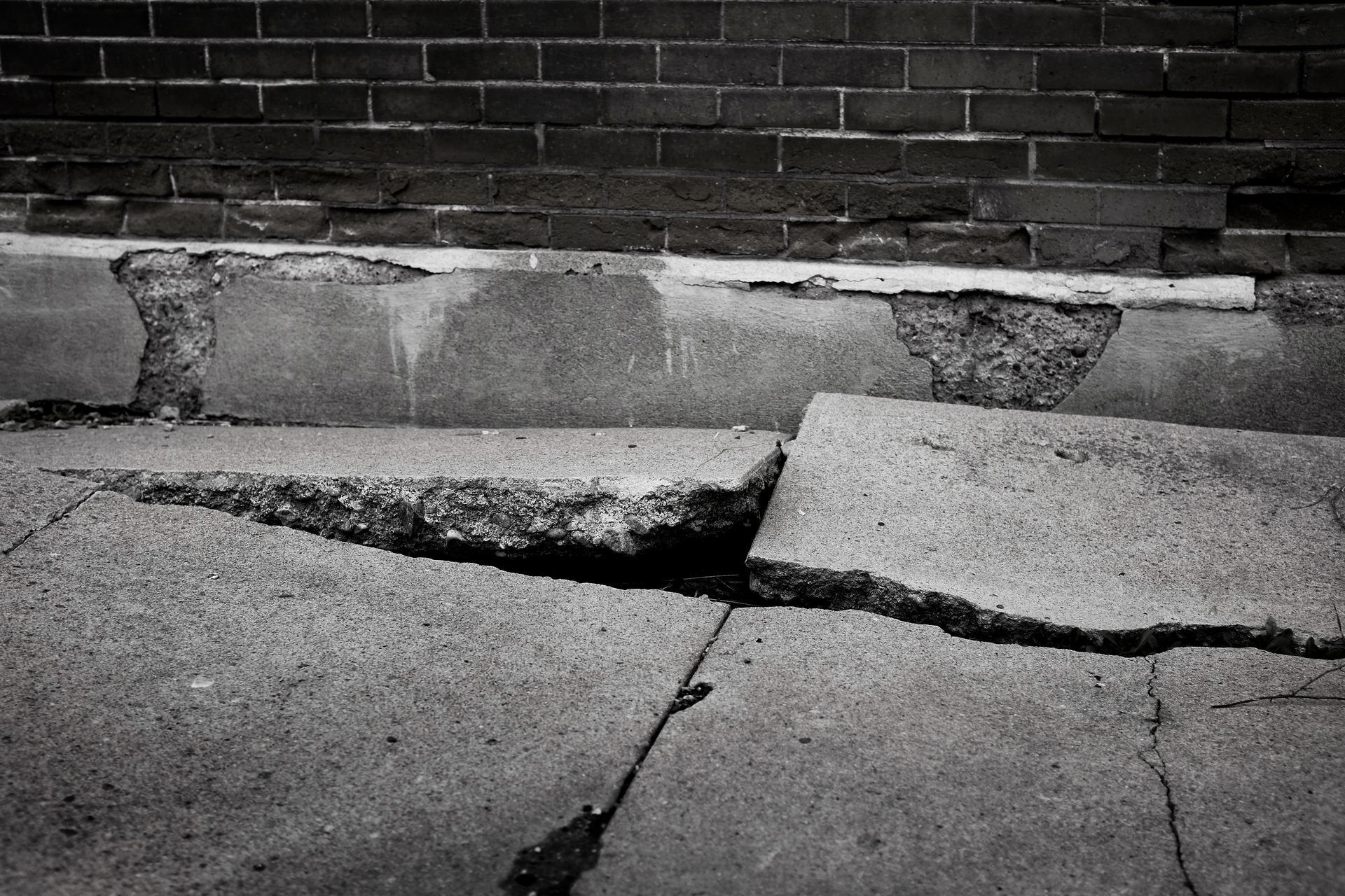 Cracked concrete slab next to a brick wall. The concrete shows significant damage and separation.