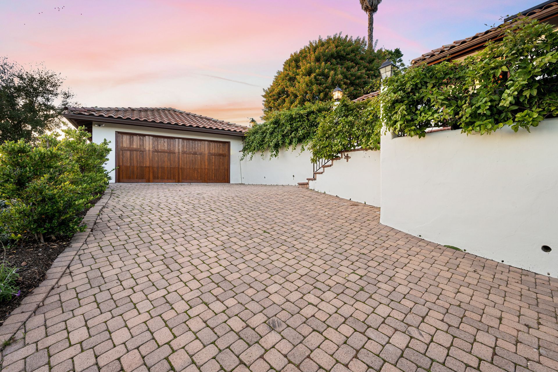 Paved driveway leading to a garage with wooden doors, framed by white walls and greenery against a pastel sky.