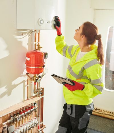 A woman in a yellow jacket is working on a boiler while holding a tablet.