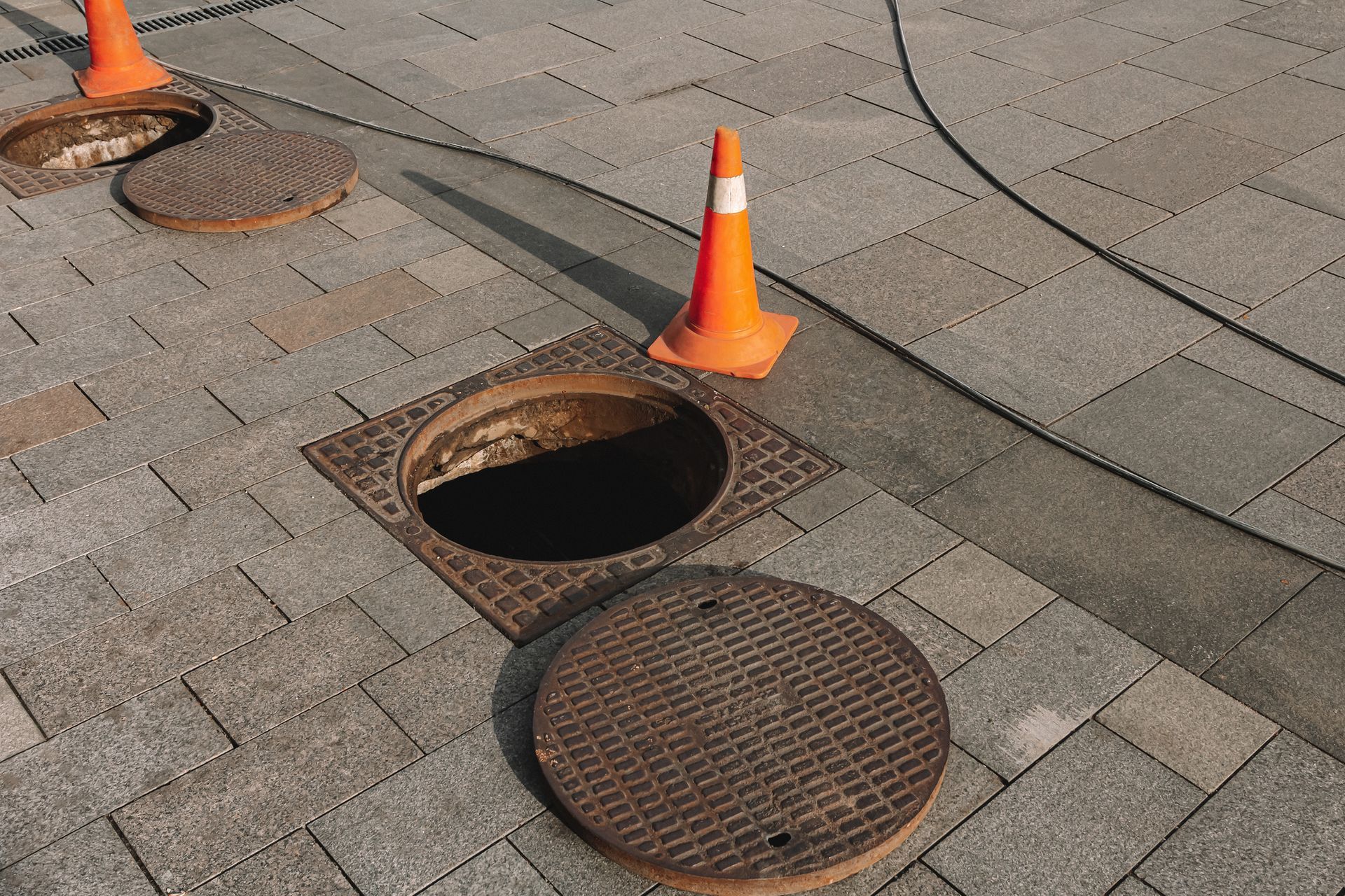 A manhole cover is open on a sidewalk next to a traffic cone.