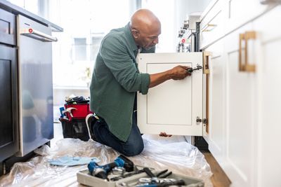A man is kneeling on the floor fixing a cabinet in a kitchen.
