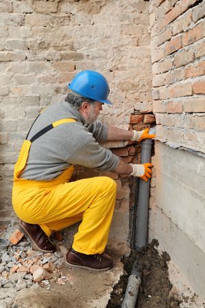 A man in yellow overalls is installing a pipe in a brick wall.